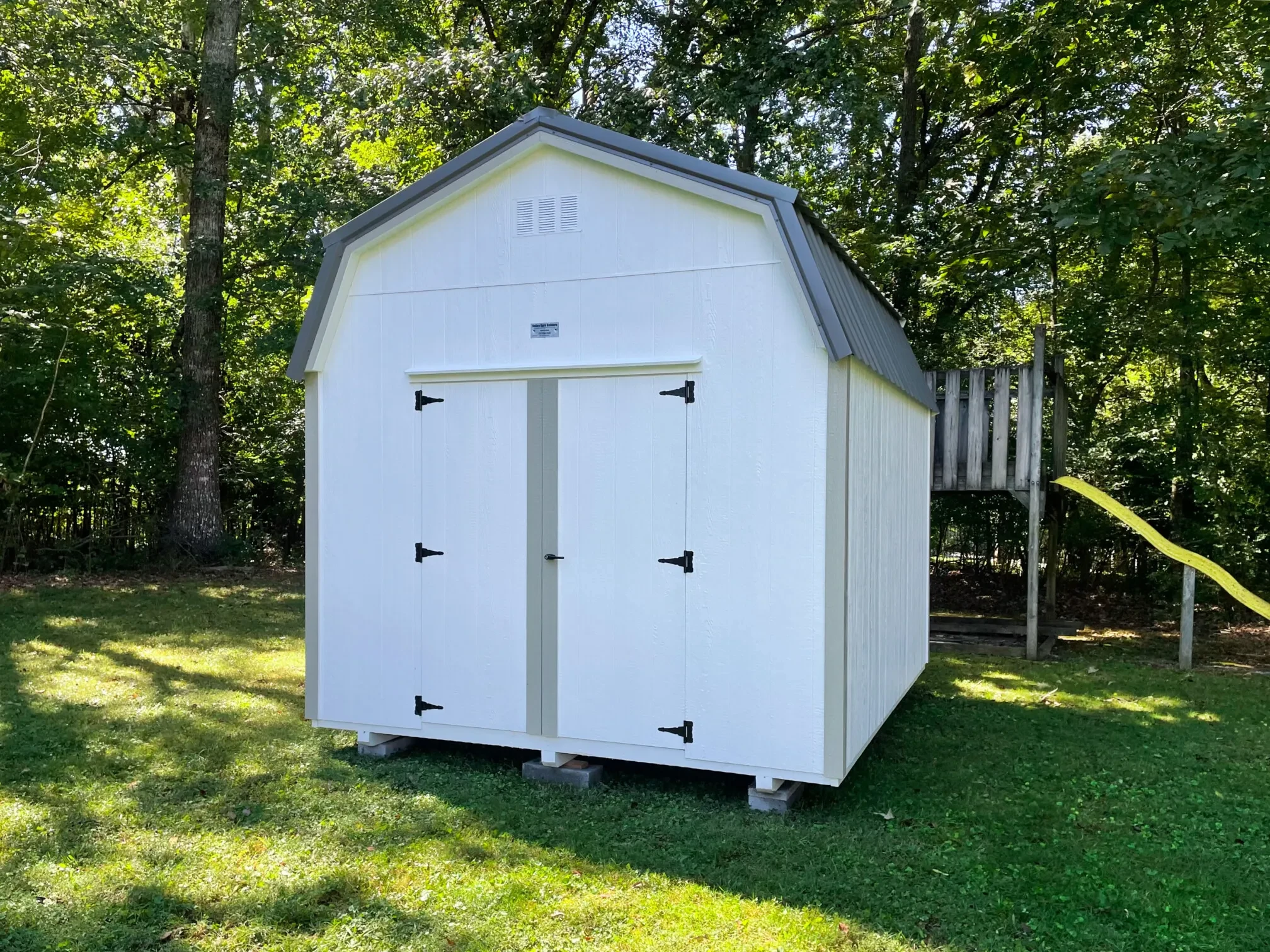 a white lofted barn sitting in a lush backyard by a playset