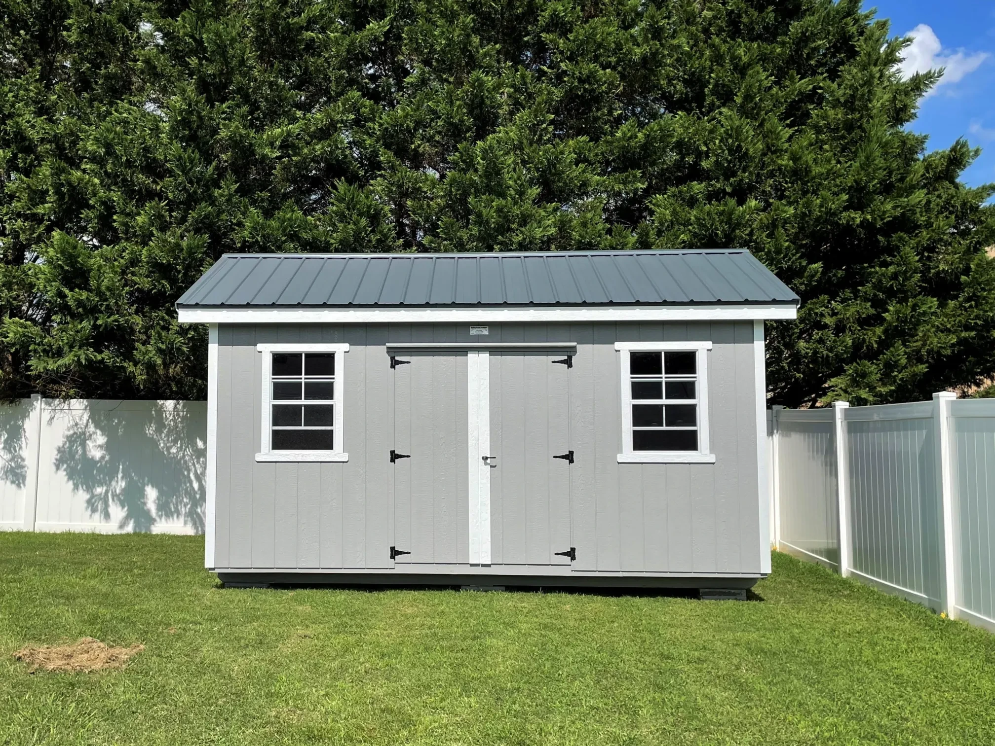 A light gray shed with windows and double doors in a backyard with a clean white fence