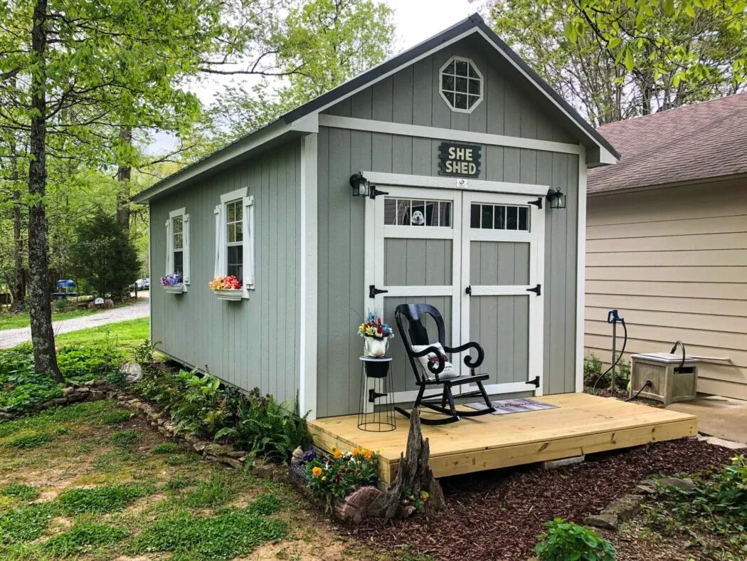 An elegantly landscaped she-shed with a porch, colorful window boxes and double doors with transom windows.