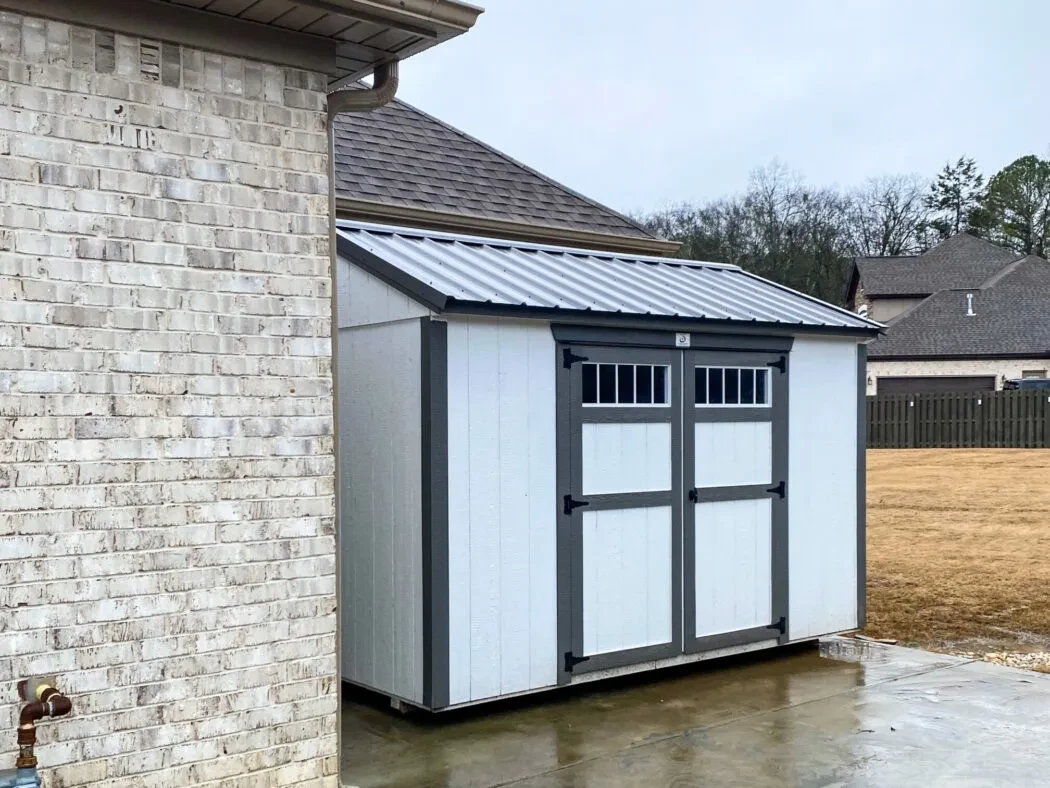 a small white shed with black roof and trim tucked next to a white brick house