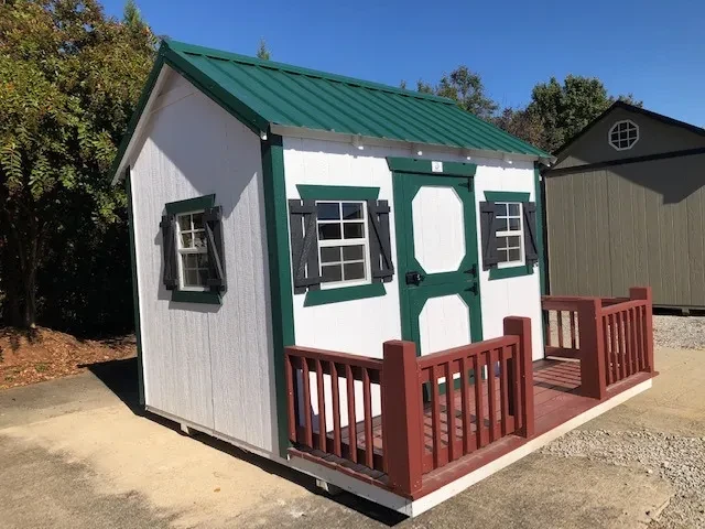 a white playhouse with green trim and a green metal roof. It also has a small porch for kids to be on