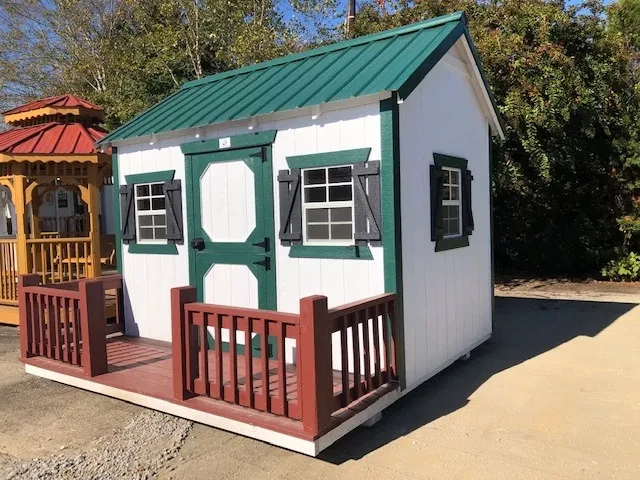 a wood playhouse with a small porch. It is white and has green trim and a green metal roof.