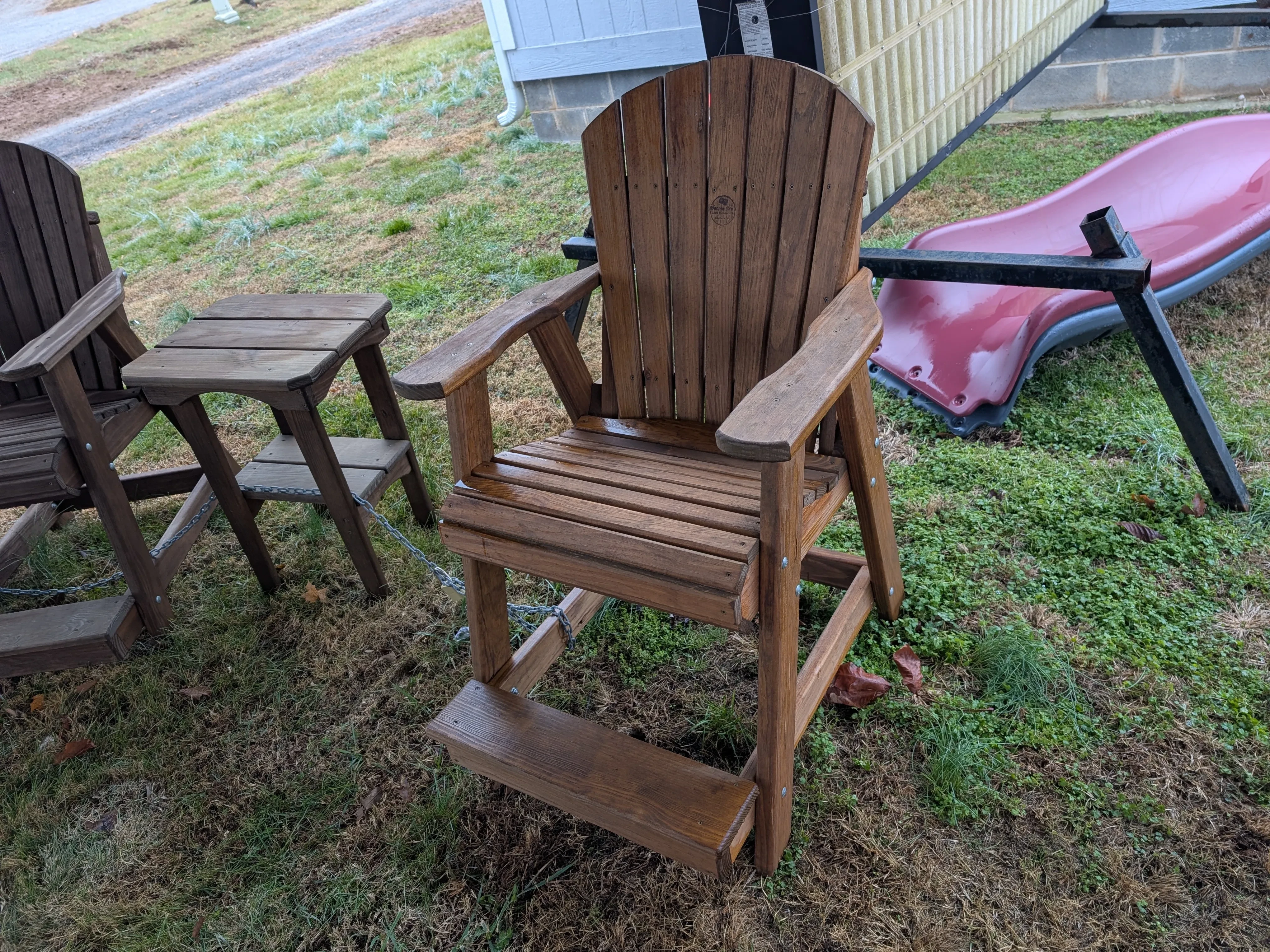 a wood stained chair in the grass next to other furniture. It has armrests and footrests.