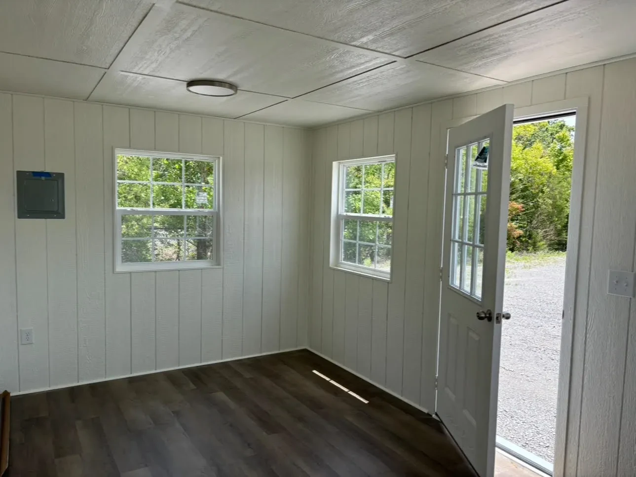 The inside of a finished out shed showing a sub-panel box next to windows and an open walk-in door.