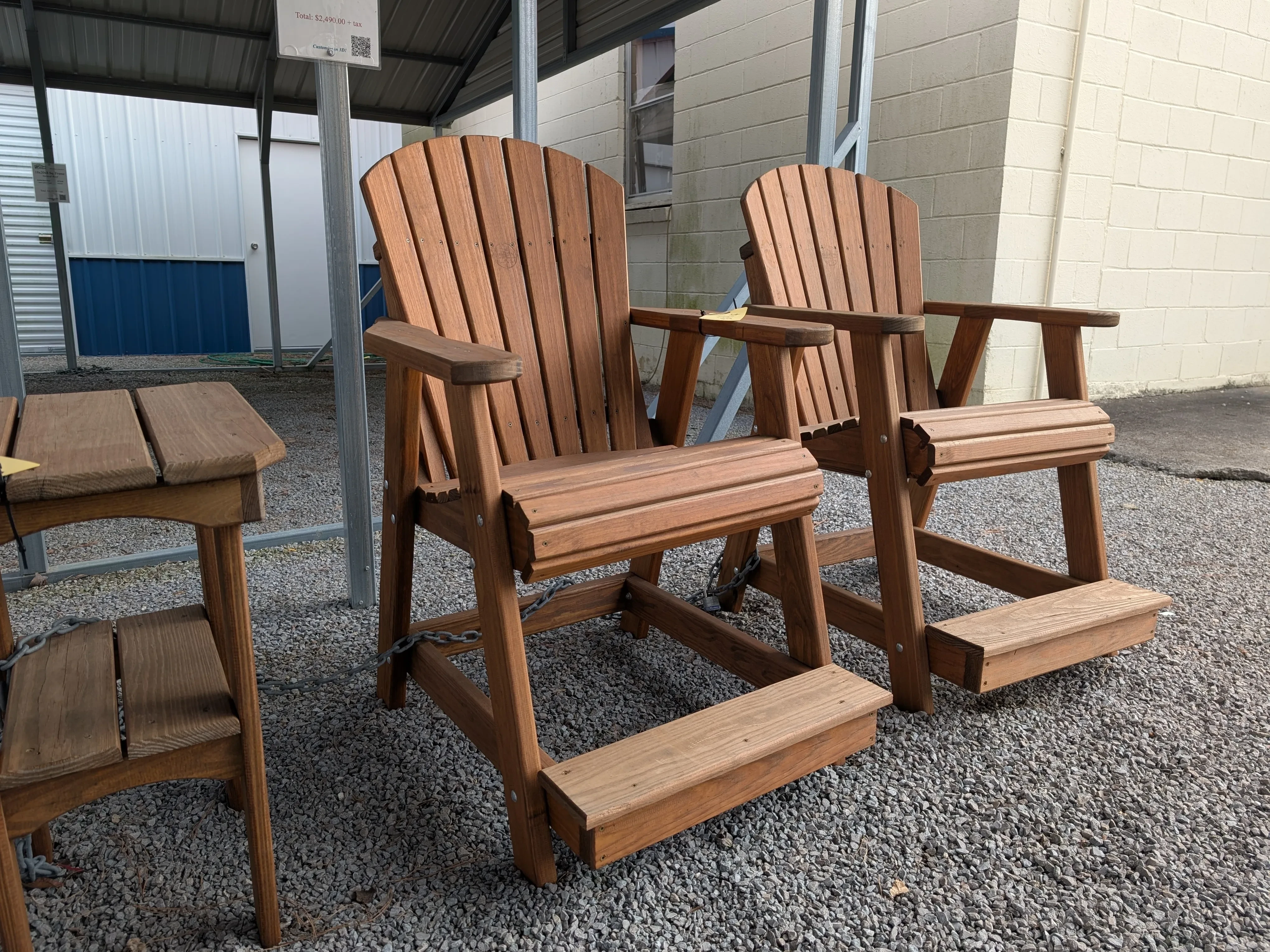 a wood stained and sealed chair right next to another chair. It has a foot rest and is sitting in the gravel