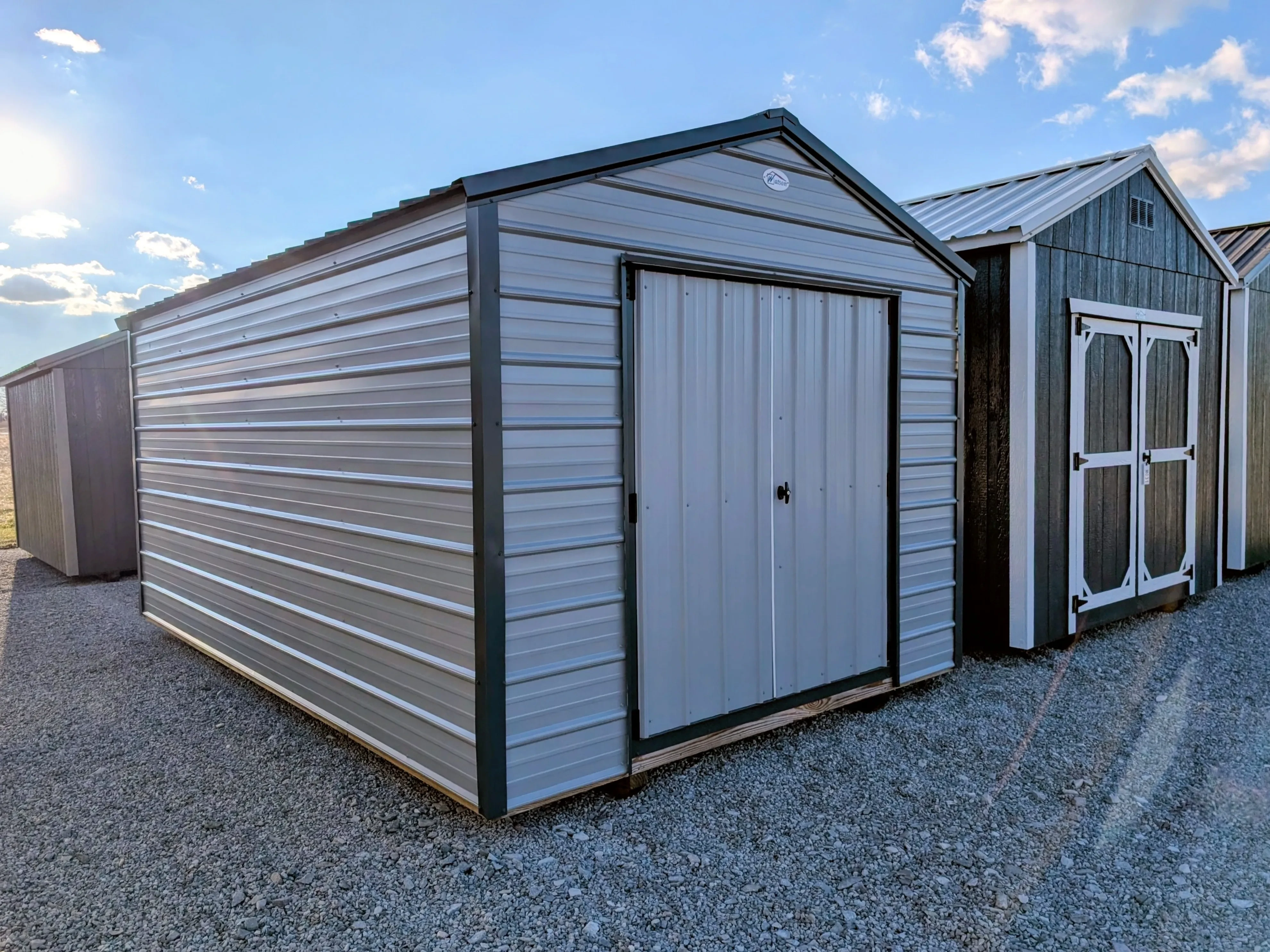 a metal utility shed next to other sheds. It has double doors and is a light gray color with dark gray trim