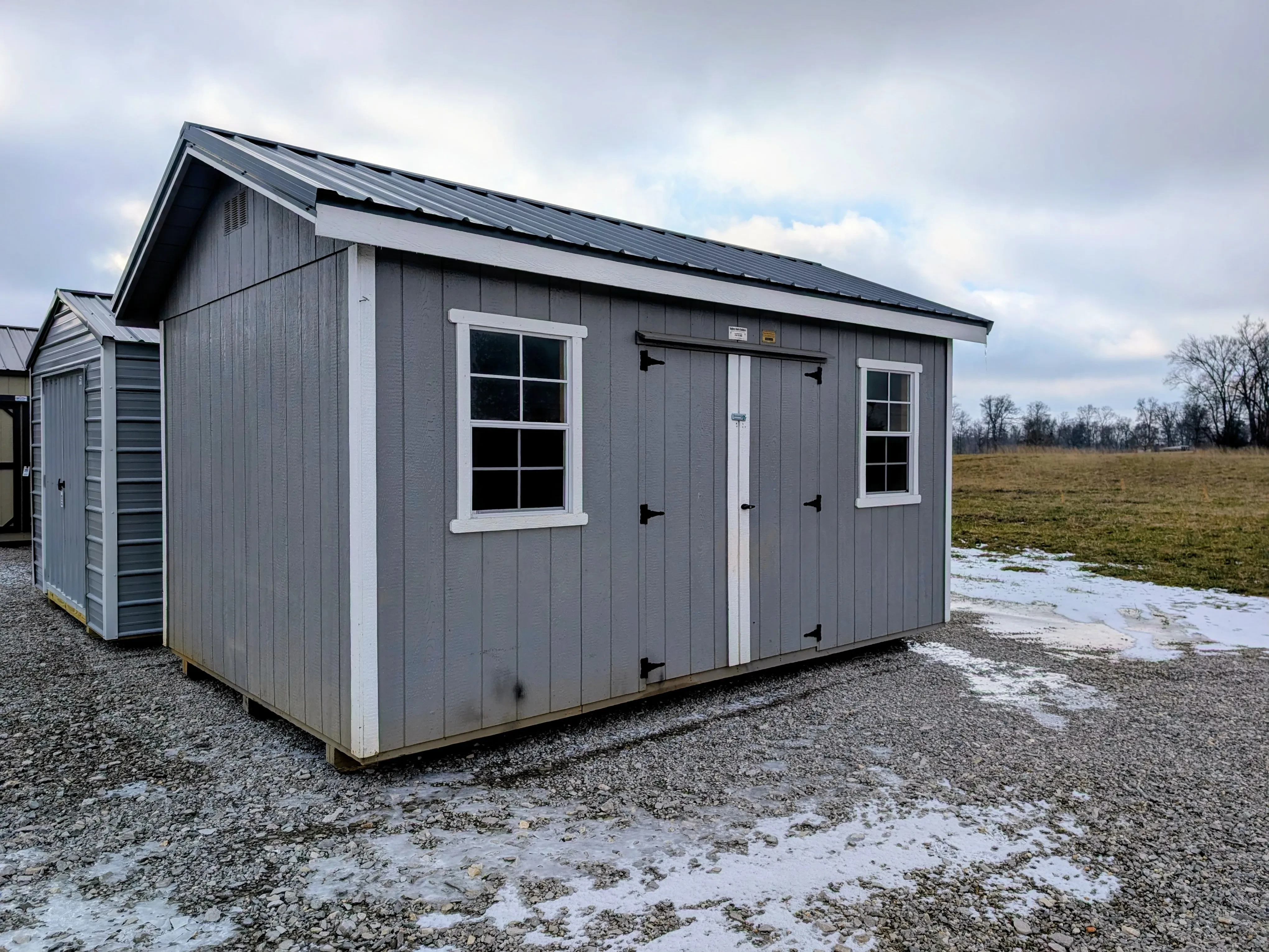 a gray shed with white trim and a roof overhang. There is snow on the ground nearby