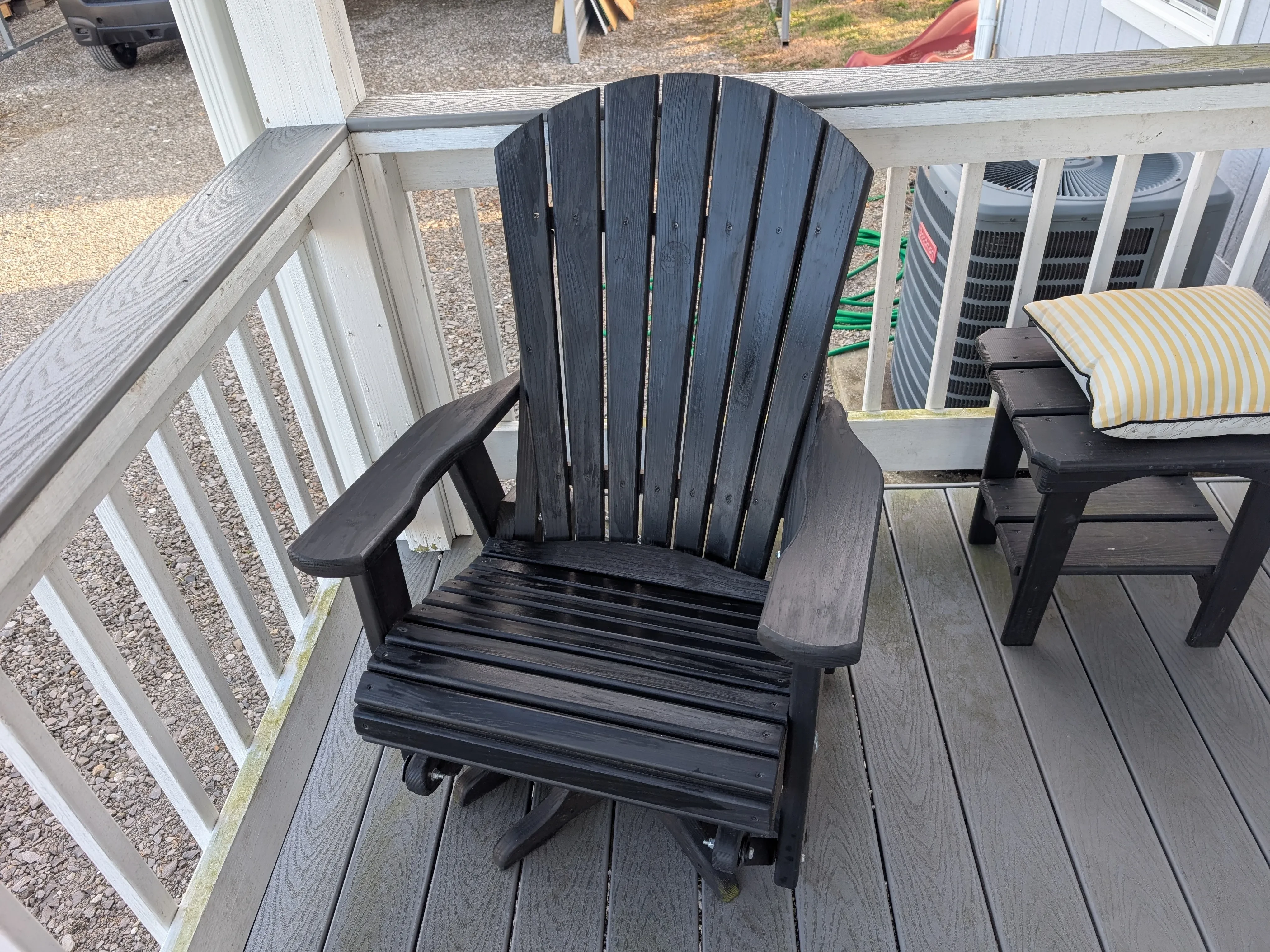 a black chair sitting on a porch. There is an AC unit behind it and a black table with a pillow on it next to it