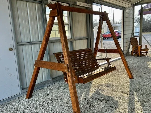 a wood swing being held up by a wood a-frame structure. It is under a carport in the gravel