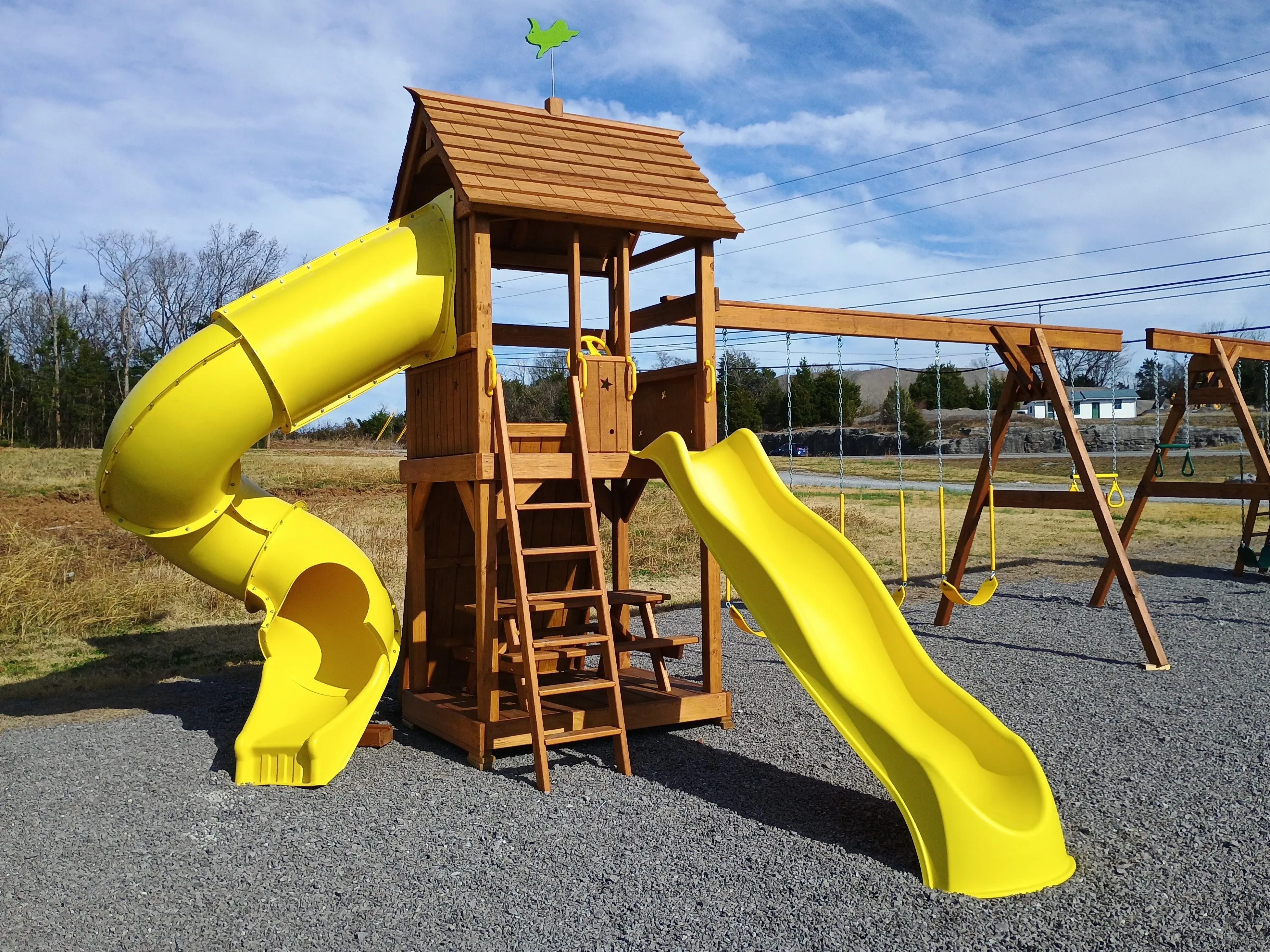 the corner view of a large playset with two yellow slides and three yellow swings. It also has a trapeze bar