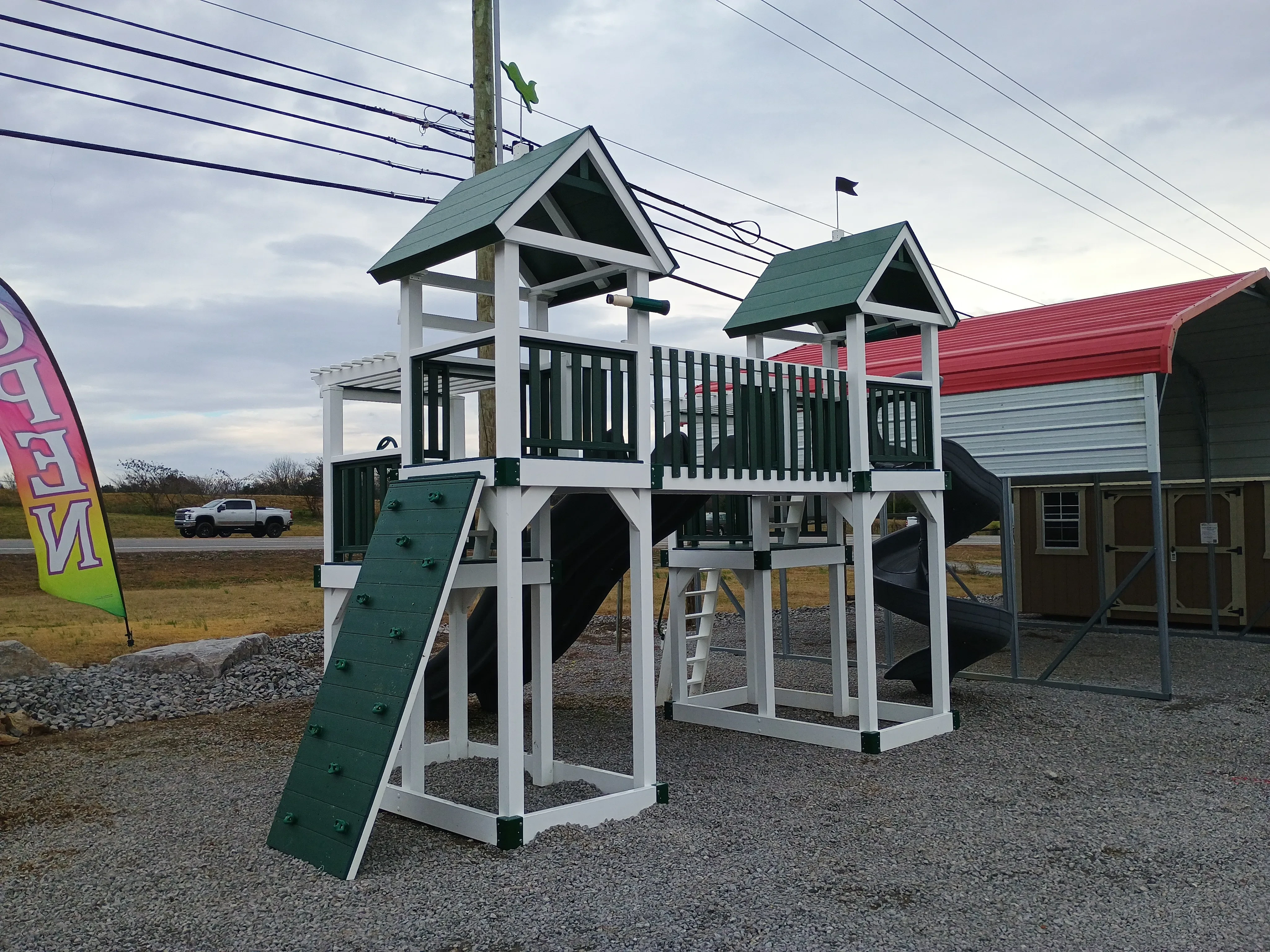 the rear view of a white vinyl playset with green trim, it has a green rockwall and is sitting next to a carport