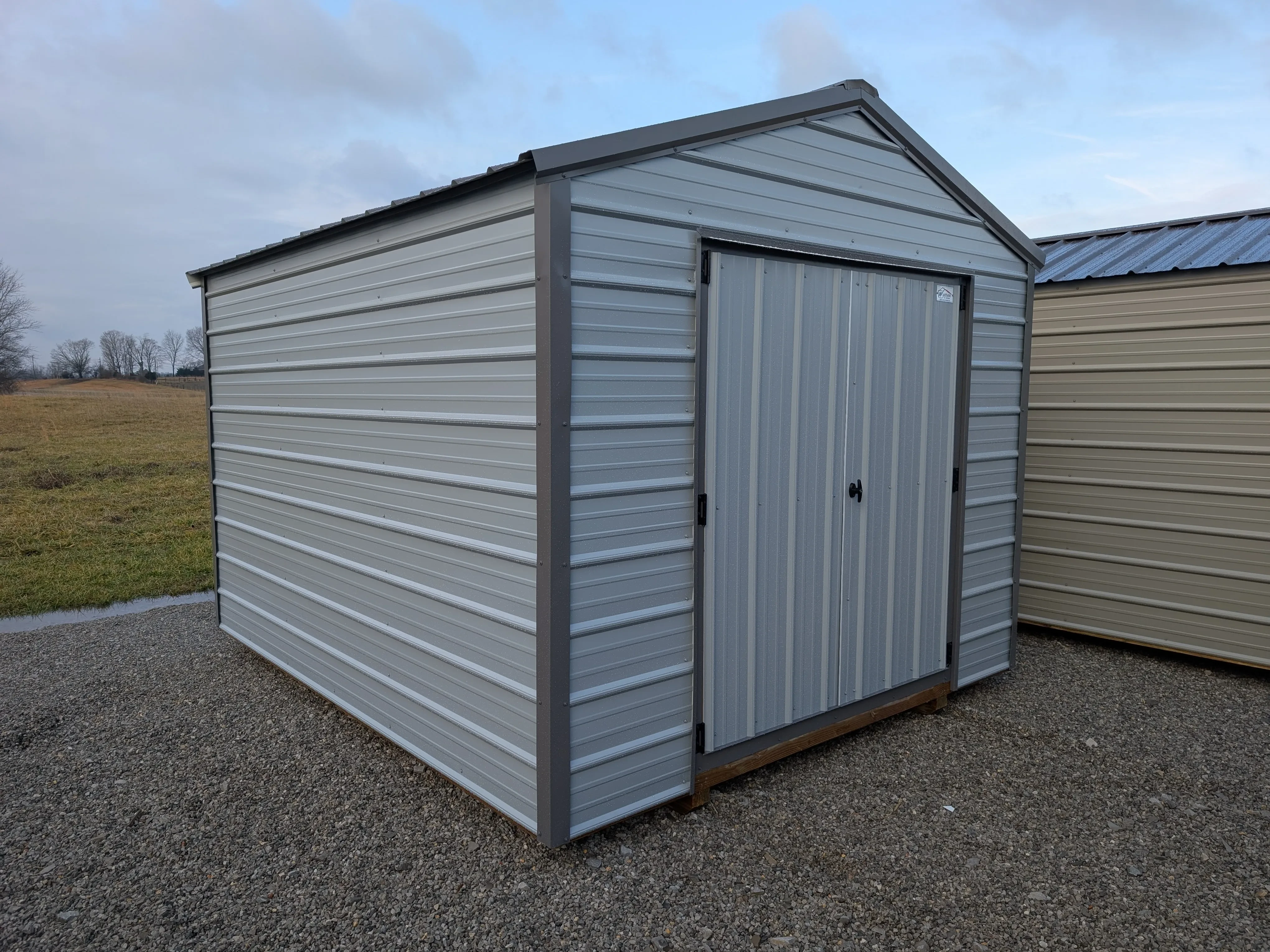a gray metal shed with double doors sitting in the gravel. It has dark gray trim