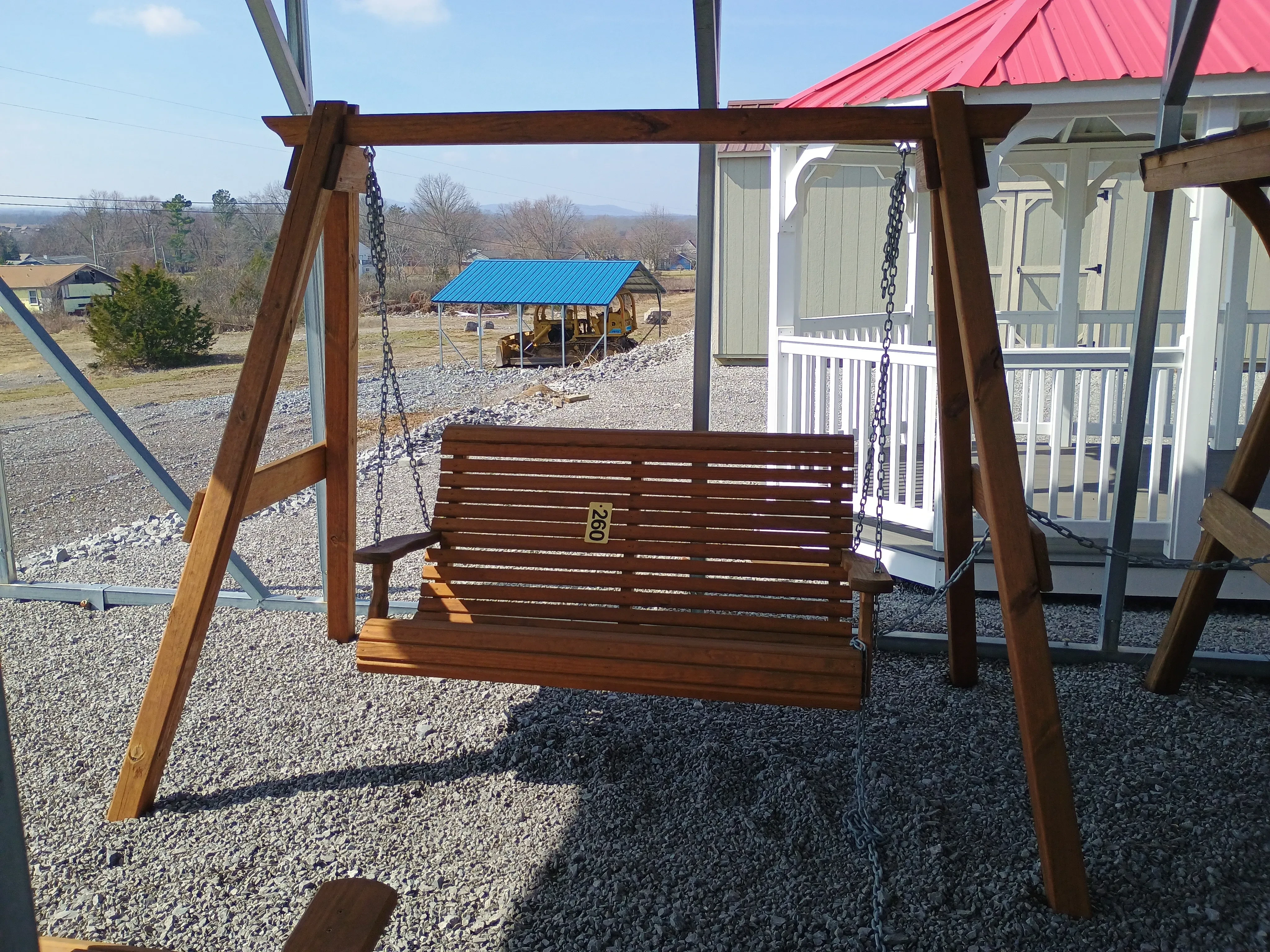 a bench attached to a wooden a-frame structure in the gravel. There is a white gazebo behind it