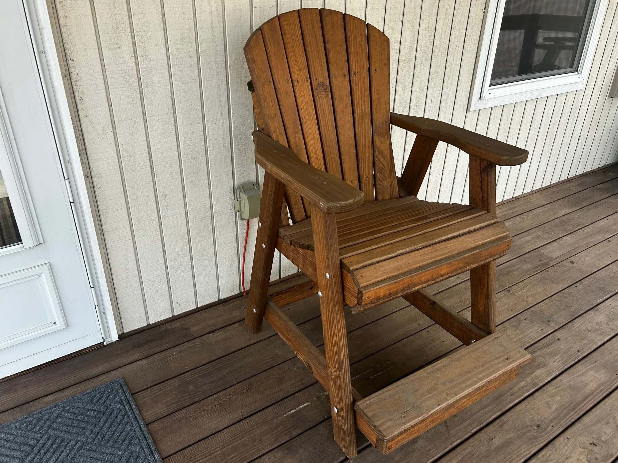 a wood stained and sealed chair sitting on a wood porch. It has armrests and footrests