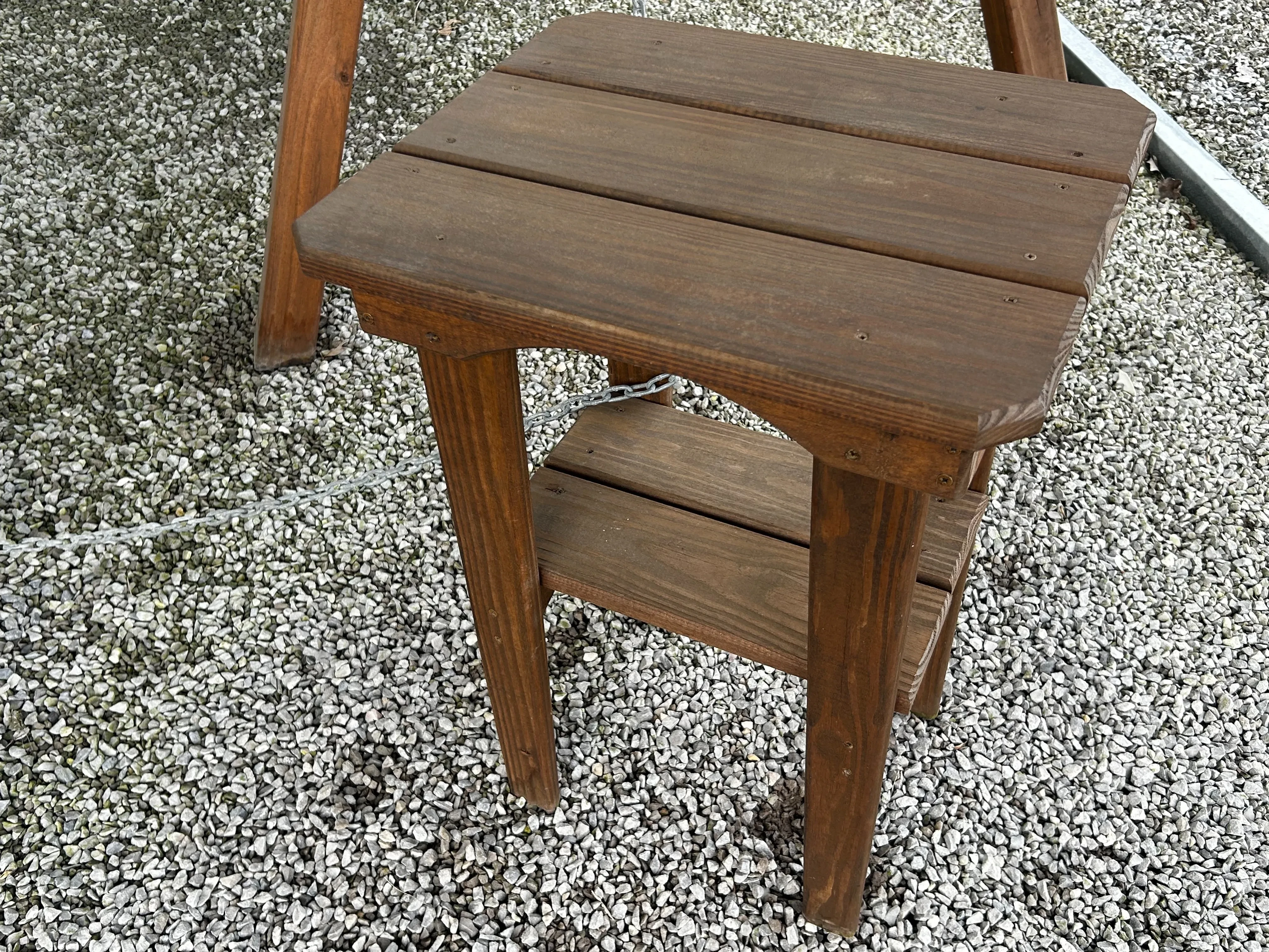 a wood table sitting in the gravel, it has a large upper tabletop and a smaller tabletop just below.