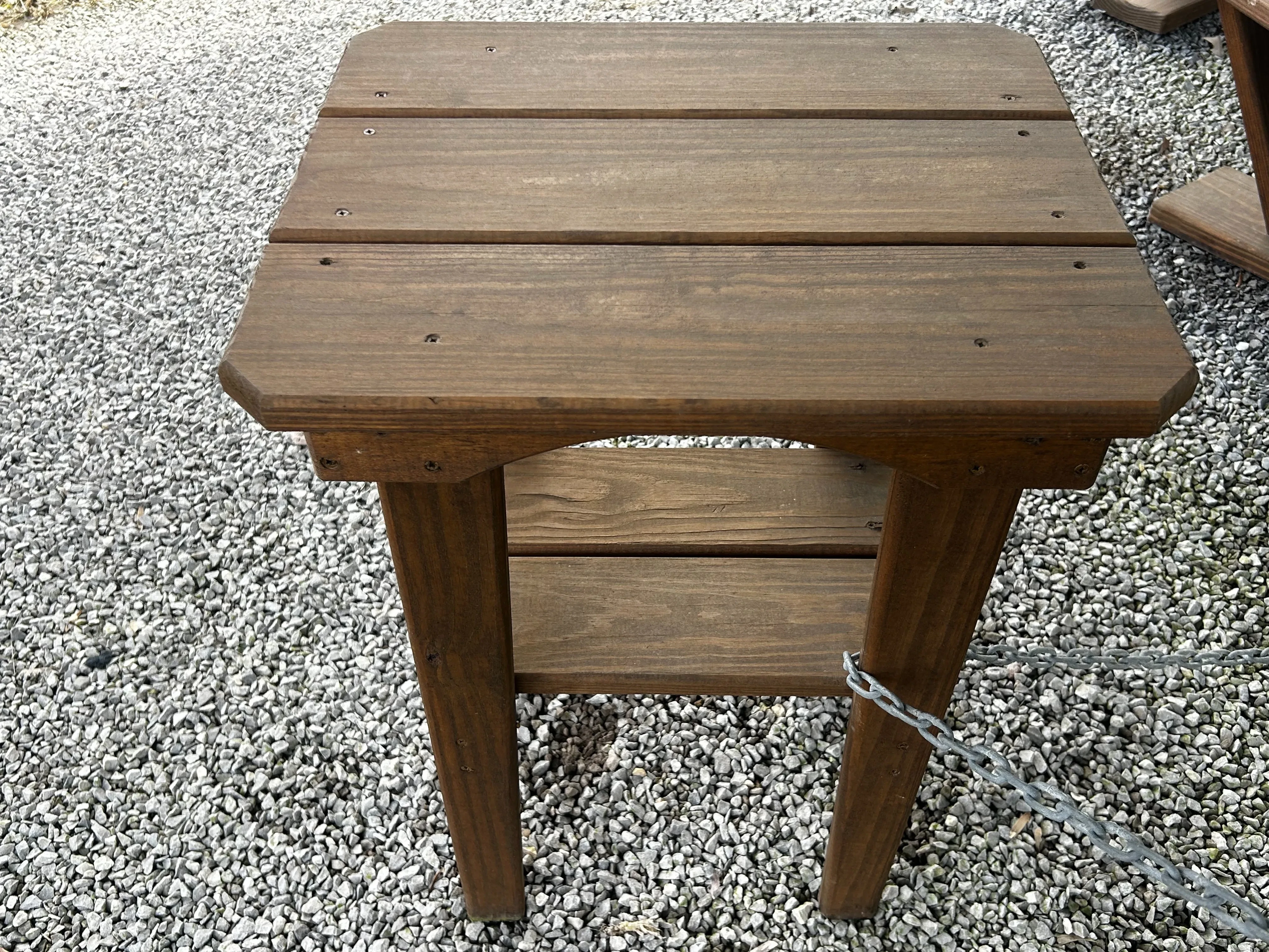 a wood stained and sealed end table sitting in the gravel. It has a chain going around one of the legs
