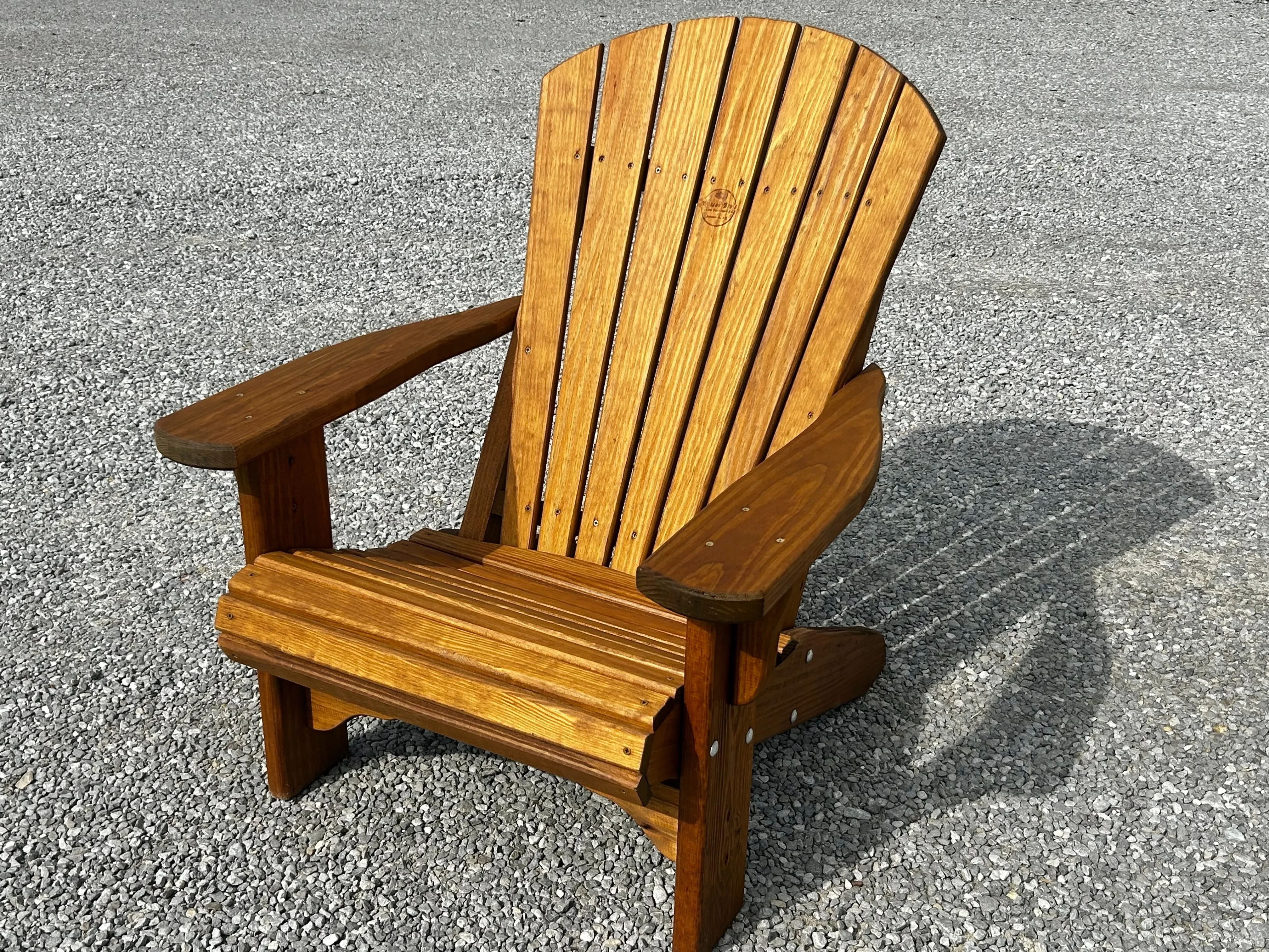 a wood stained and sealed chair sitting in the gravel. It's shadow is being casted behind it. It has arm rests