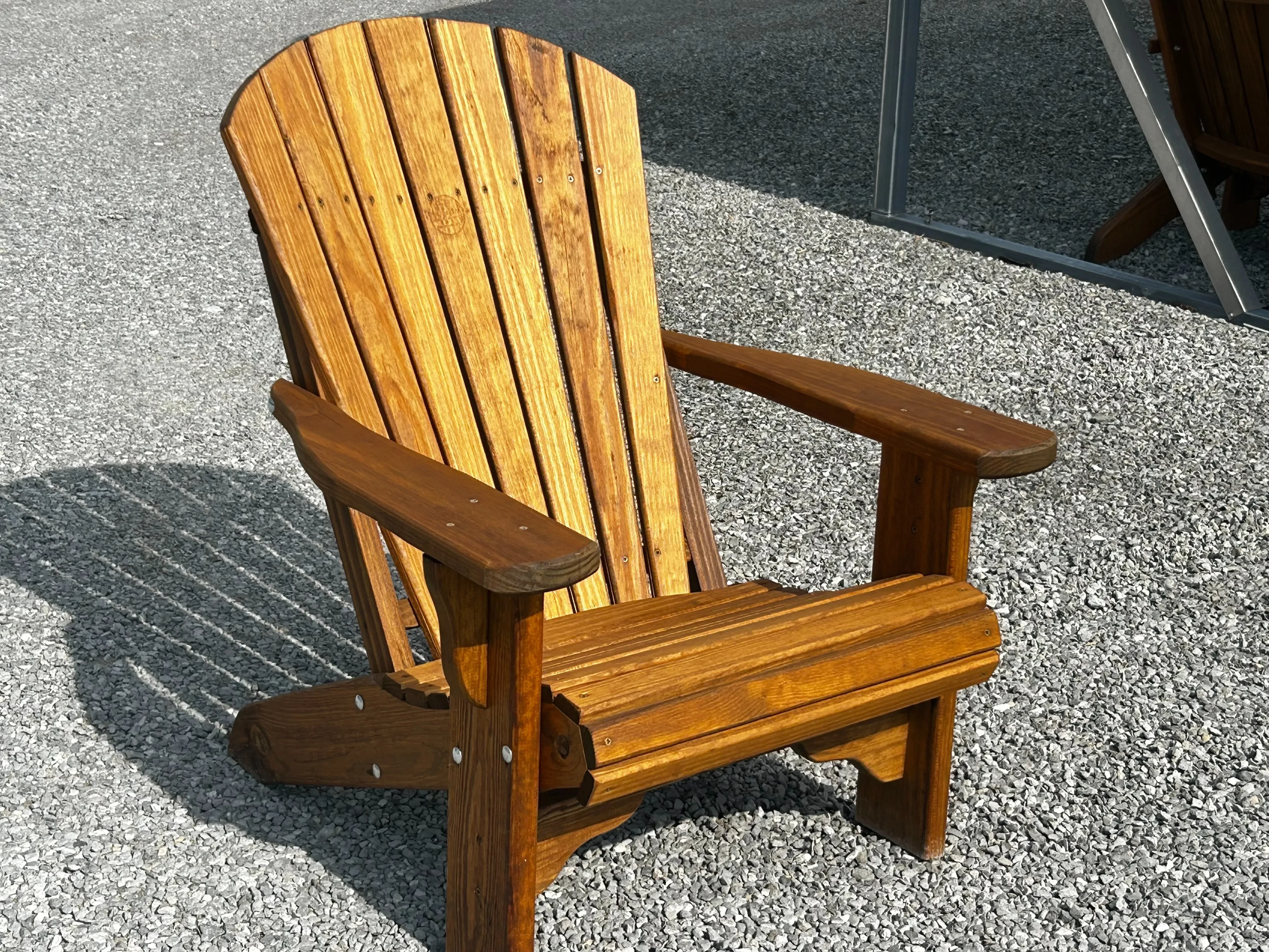 a wood stained and sealed chair sitting in the gravel. It has a shadow behind it. You can see a carport in the background