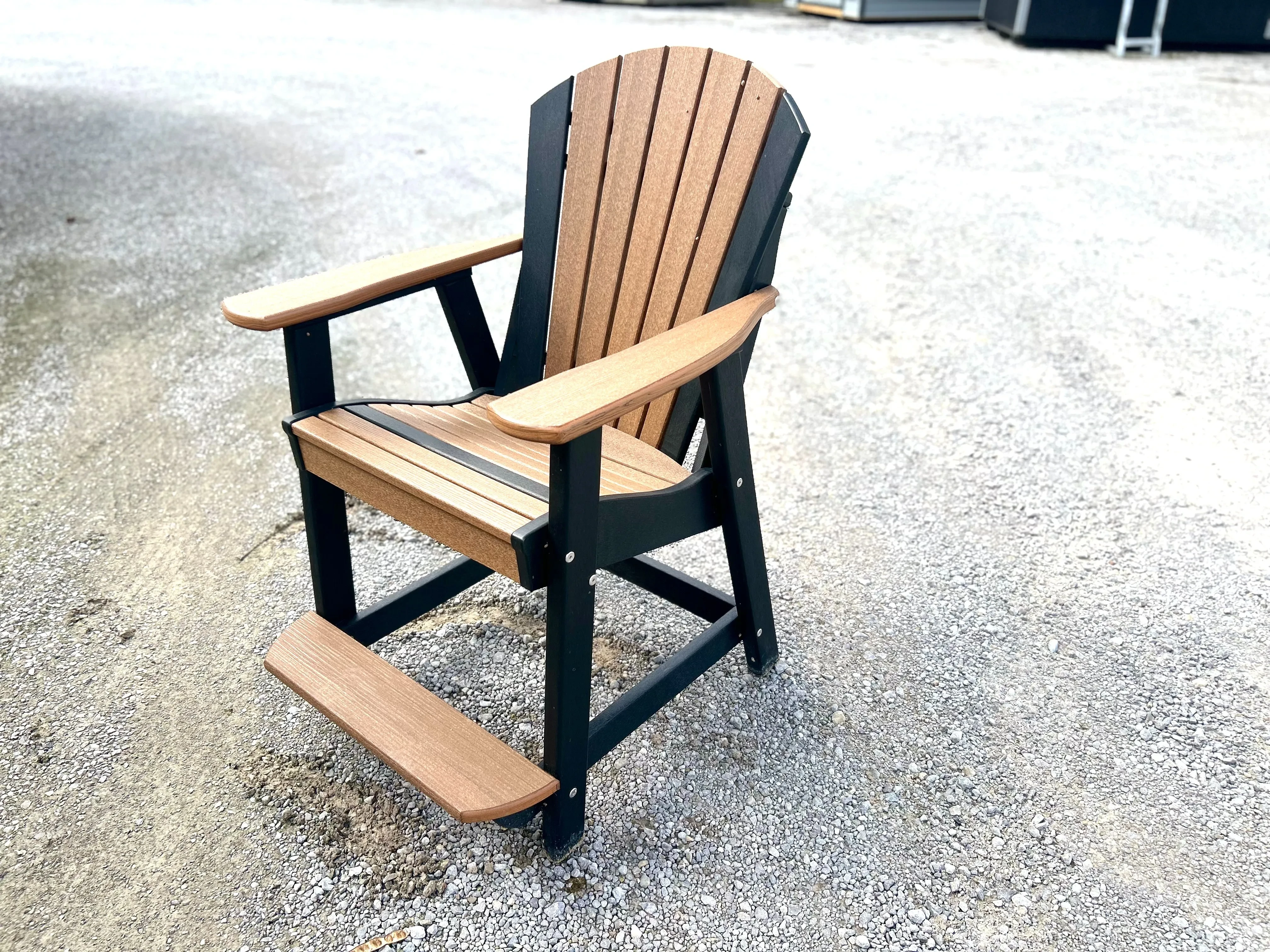 a brown chair with black accents sitting in the gravel. It has armrests and footrests. It is made out of polywood