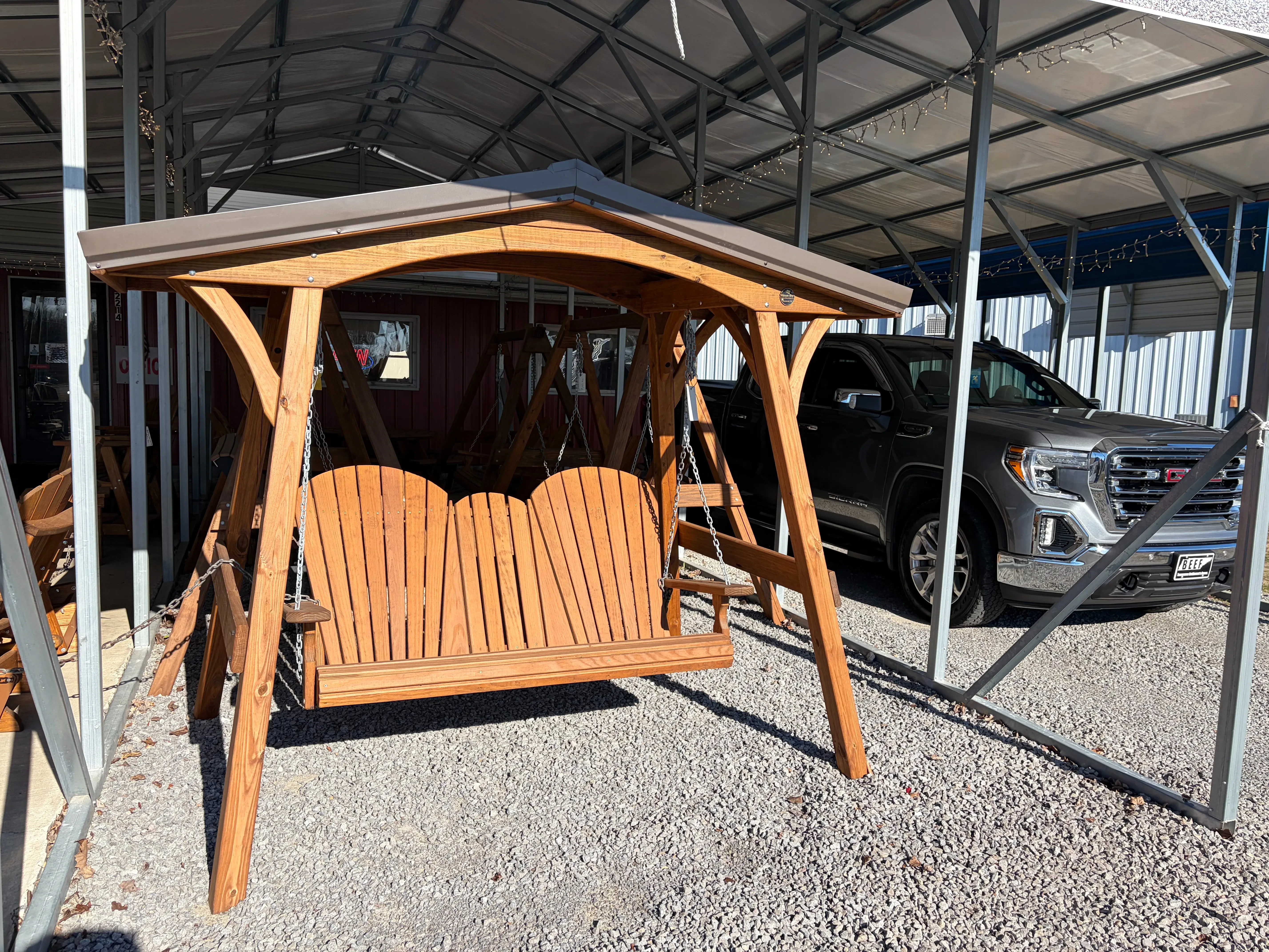 a wood bench being held up by a wooden a-frame structure with a metal roof on top. It is sitting in the gravel.