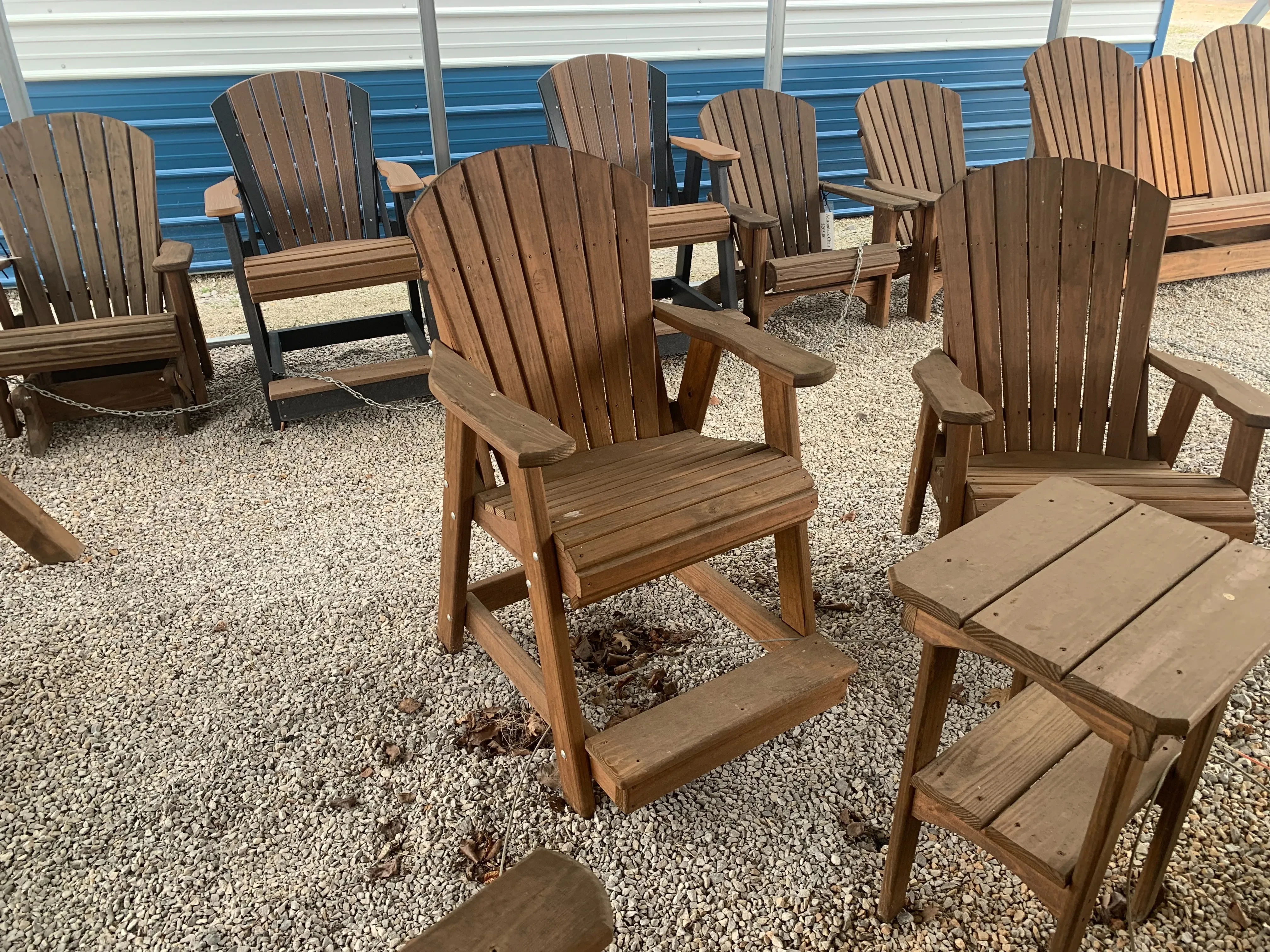 a nicely stained and sealed chair sitting in the gravel. It has armrests and footrests. There is other furniture nearby
