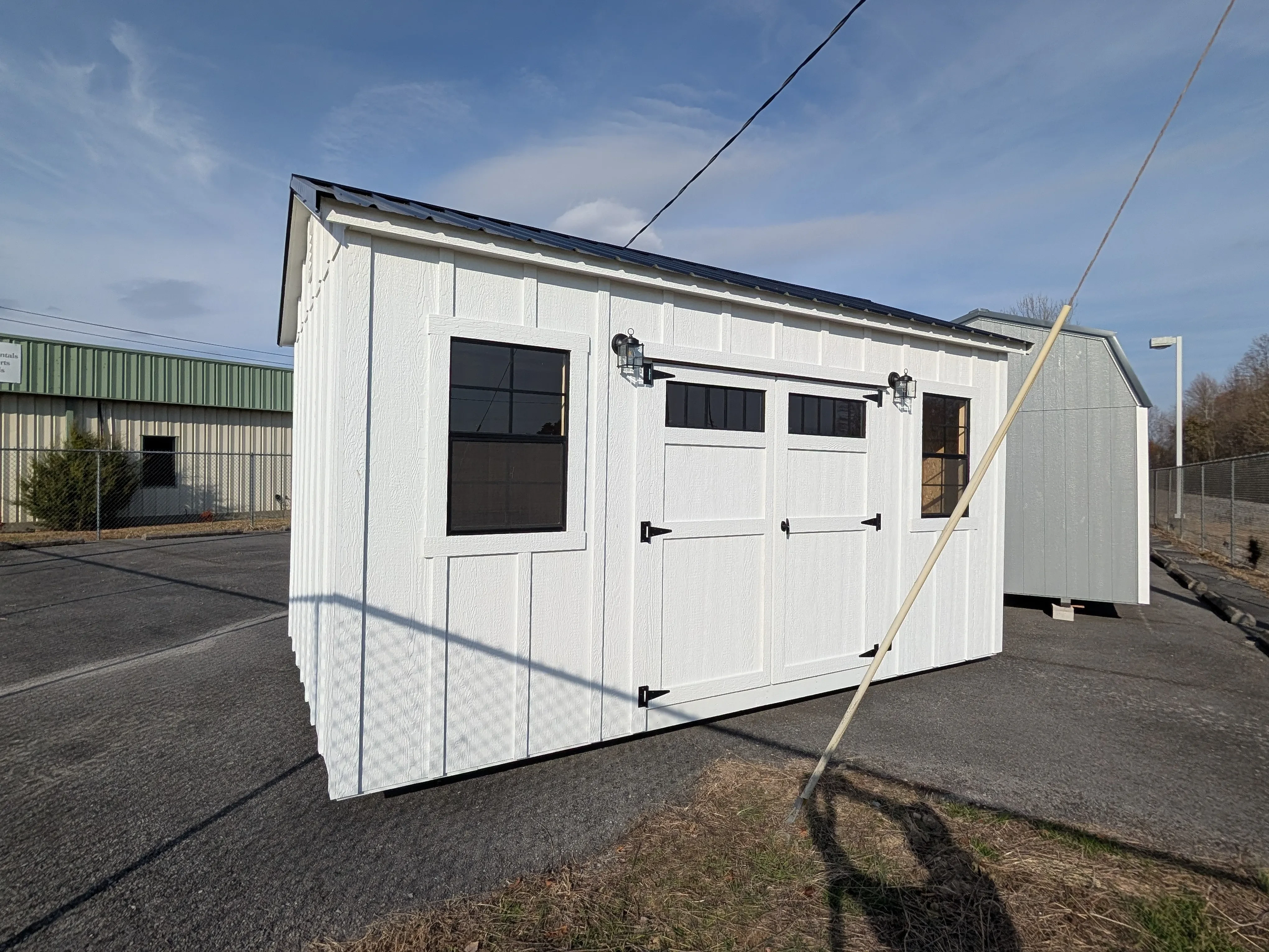 a white board & batten shed with black windows and outside lights. It has a black metal roof and is near other sheds.