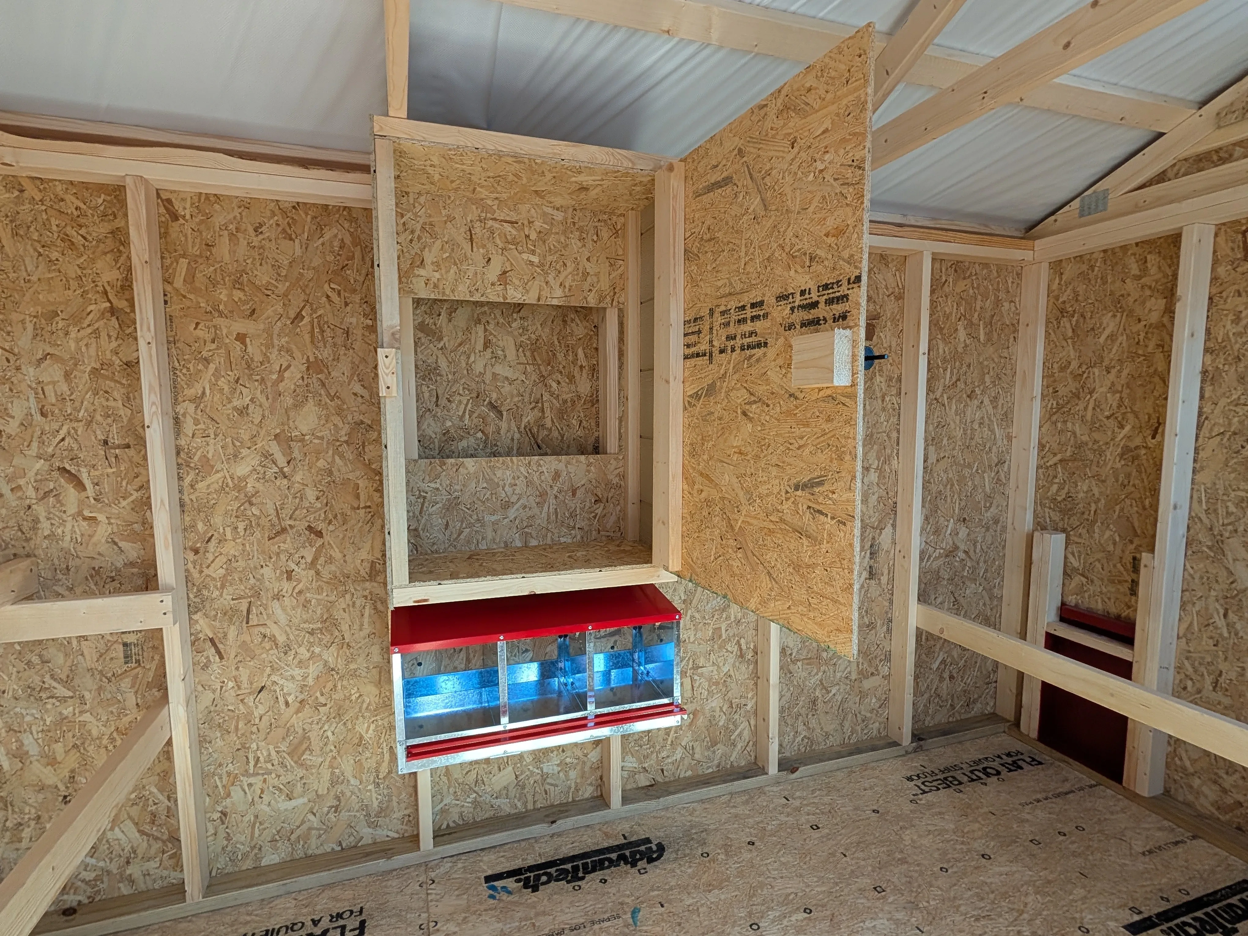 inside a wood chicken coop. It has a metal nesting box with 3 spaces and a storage cabinet above them.