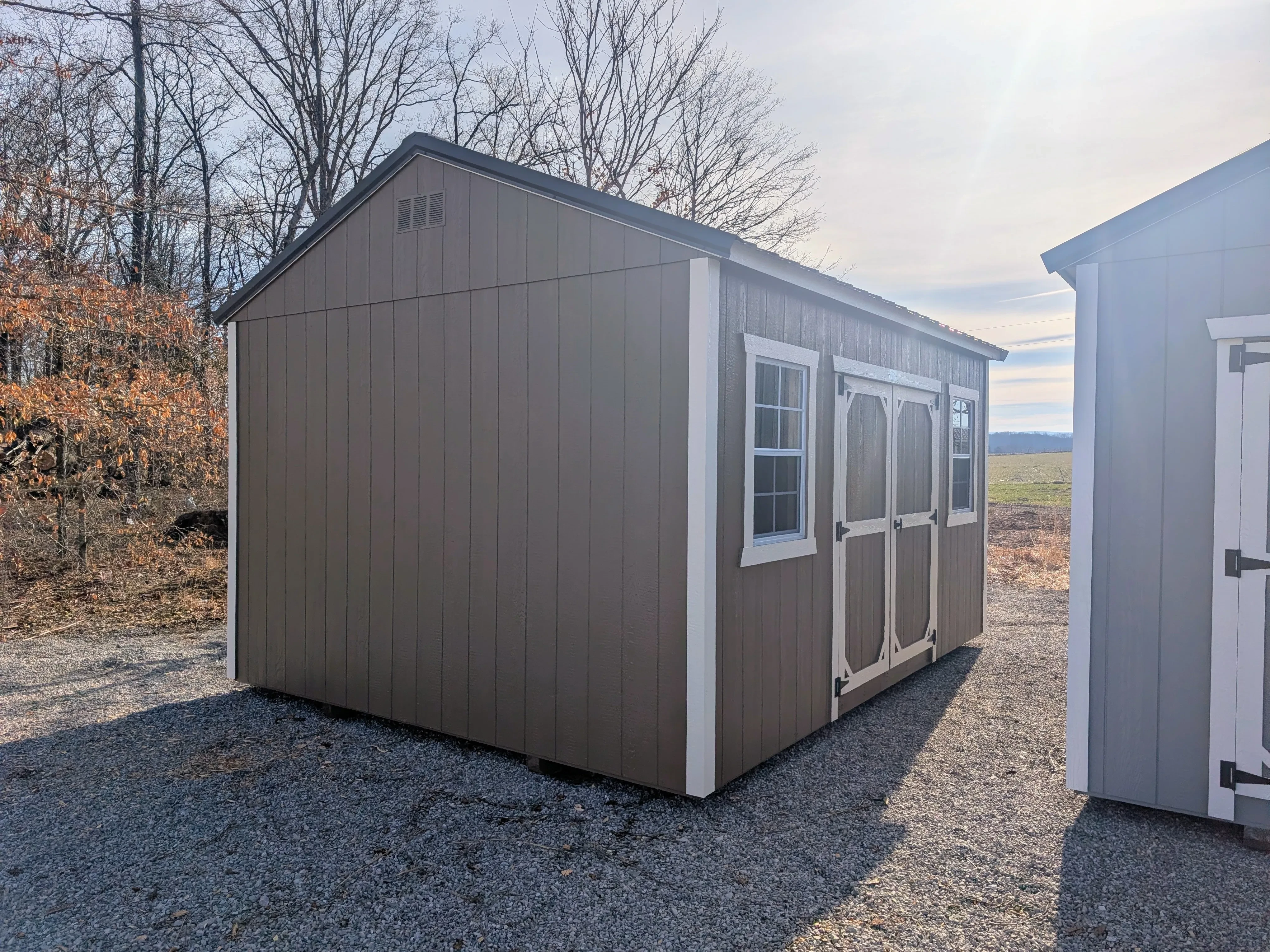 a brown garden shed with white trim and double doors. It is sitting in the gravel. It has two windows
