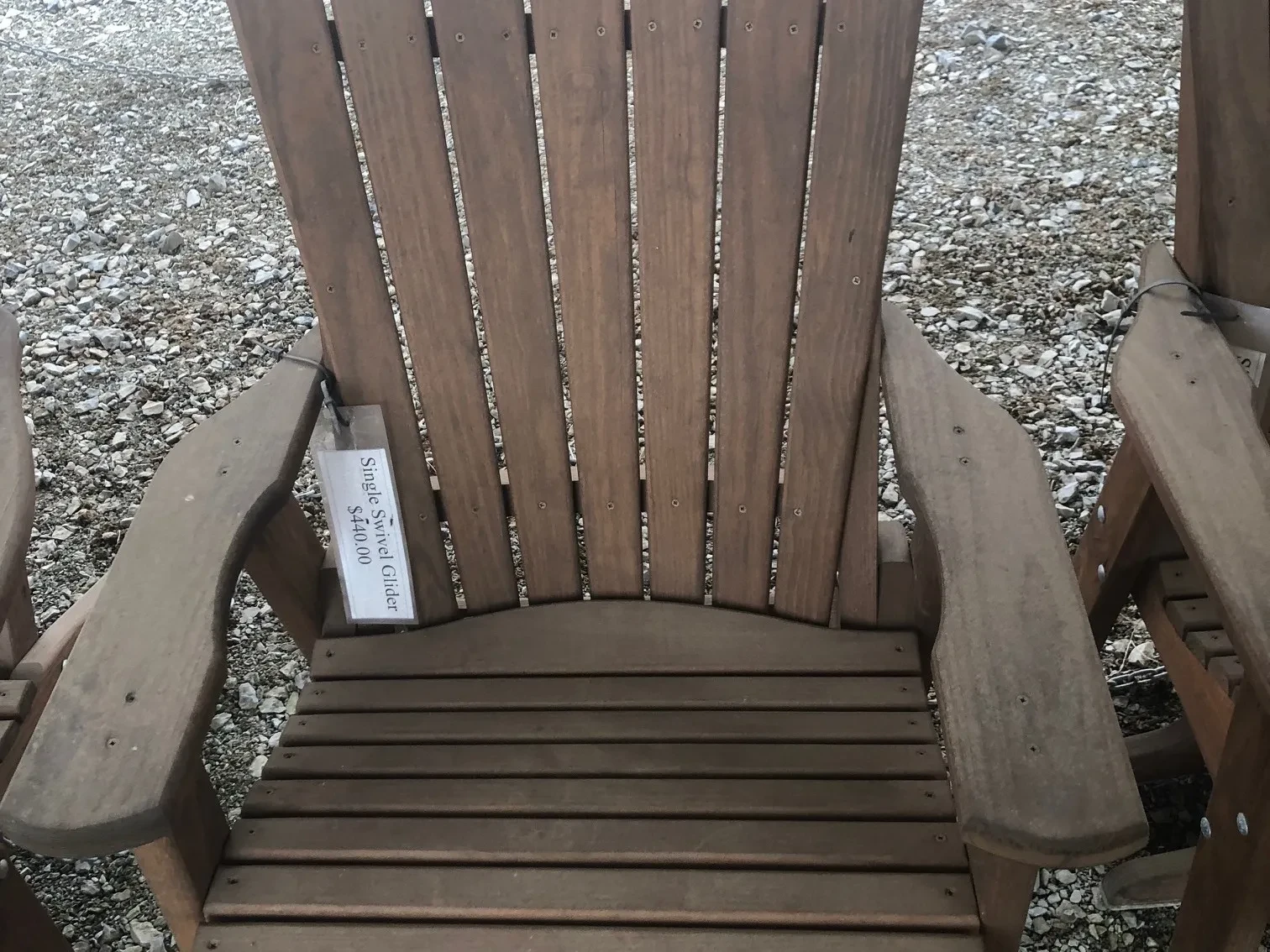 a wood stained and sealed chair sitting in the gravel. It has arm rests. It is next to other chairs