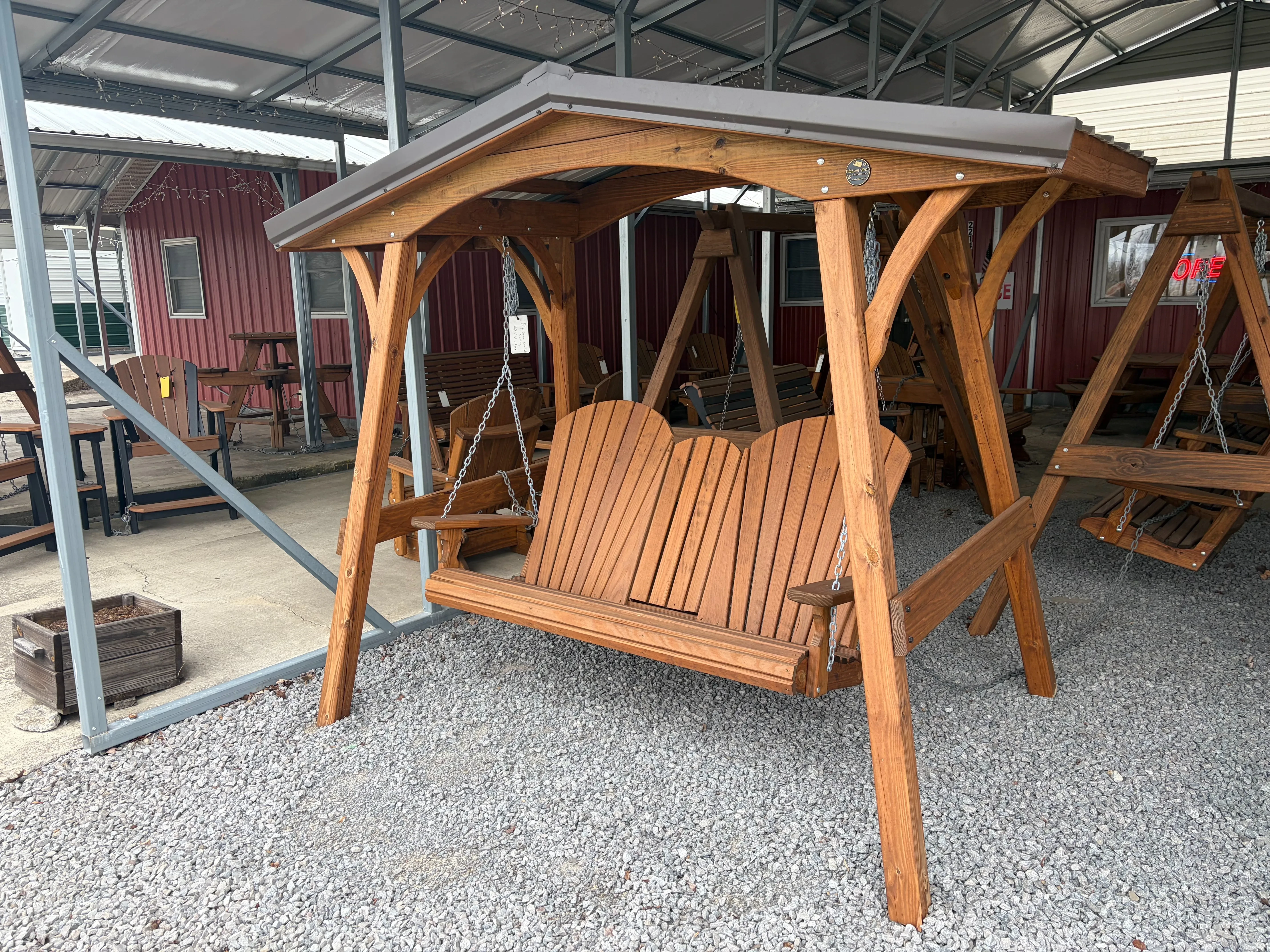 a wood bench swing attached to a wooden a-frame structure with a metal roof on top by chains. It is in the gravel