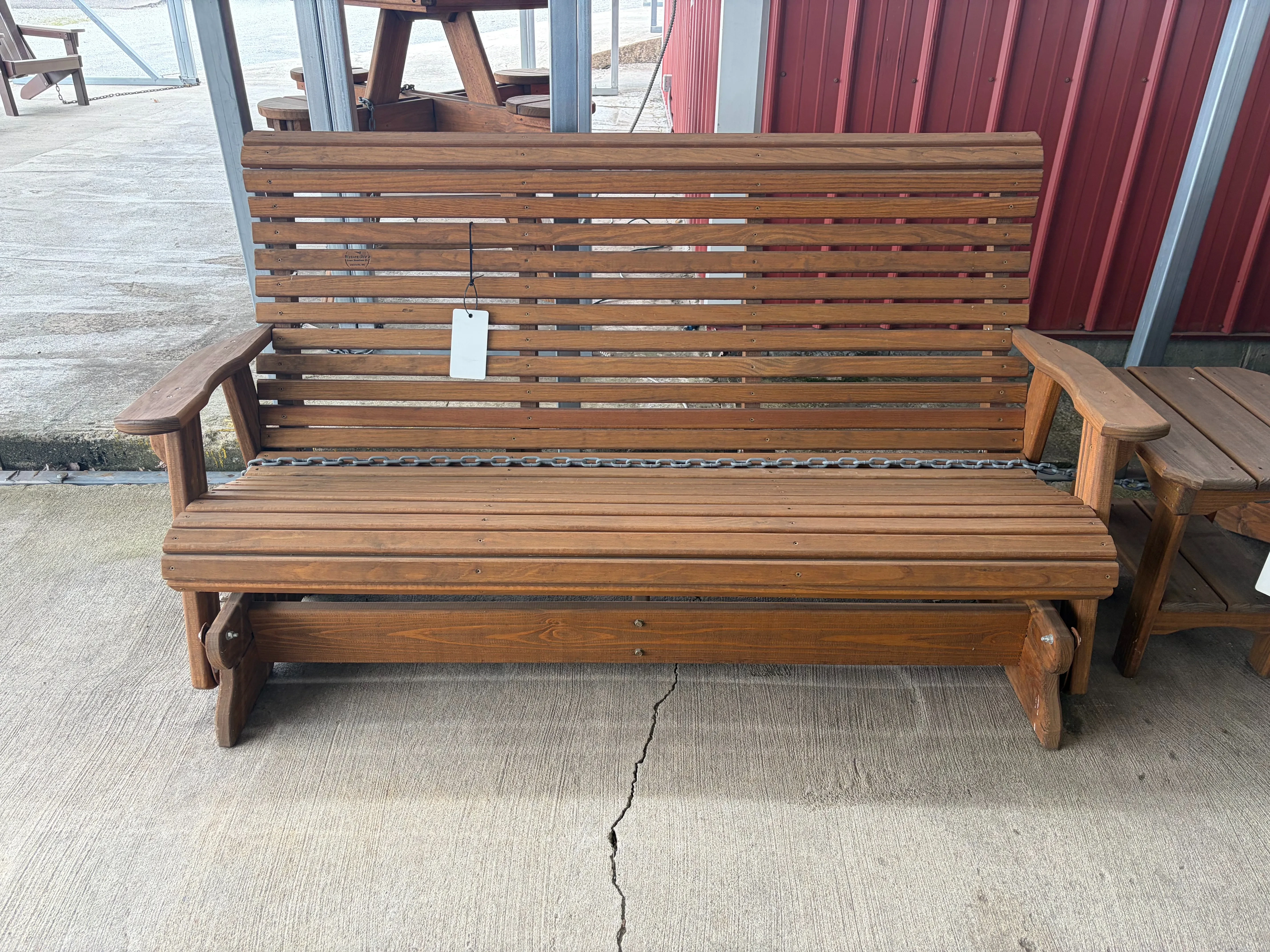 a wood stained and sealed bench sitting on concrete. It has arm rests and can glide back and forth