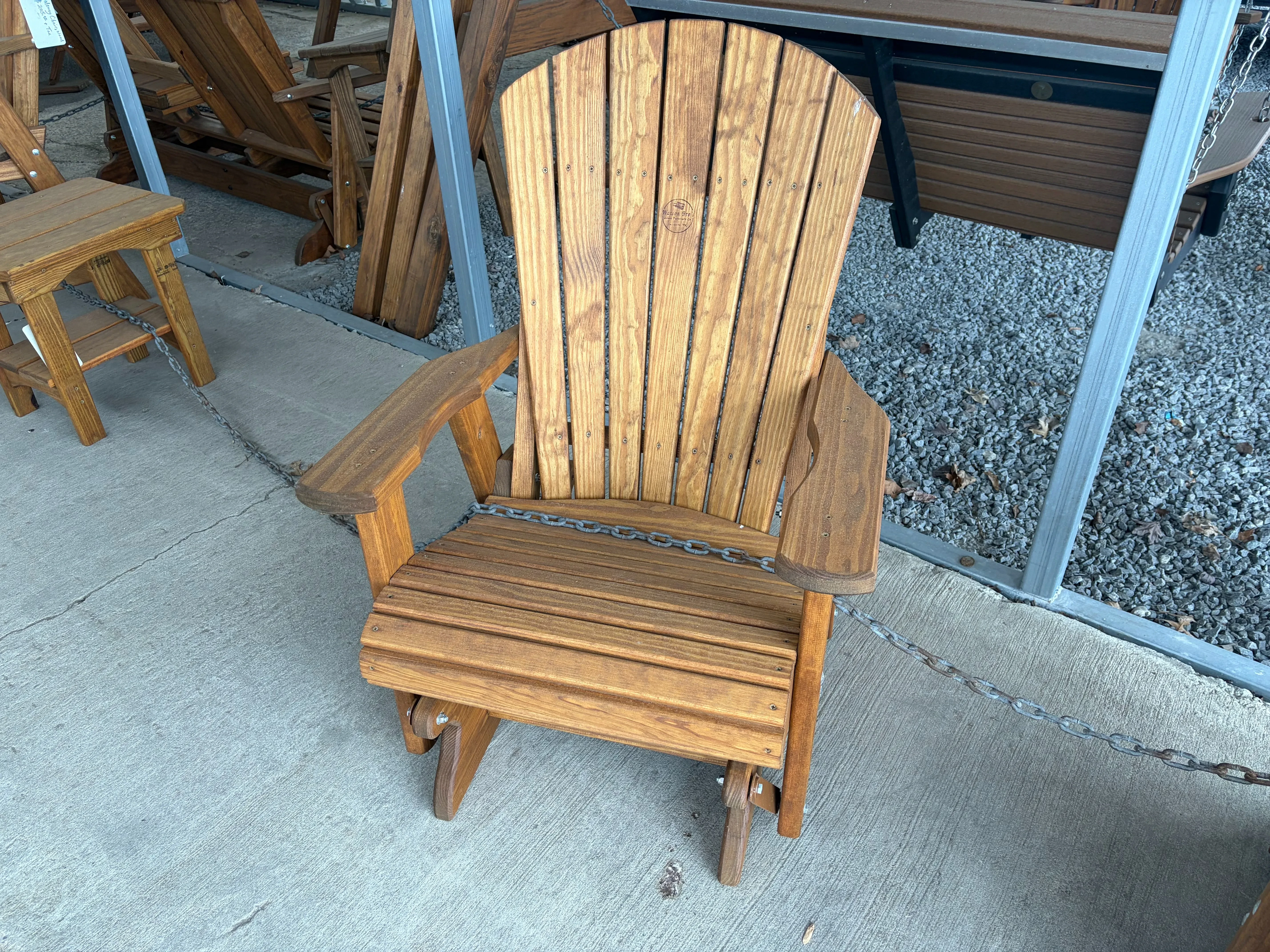 a wood stained and sealed chair sitting on concrete. It has arm rests. It is chained up to other furniture nearby.
