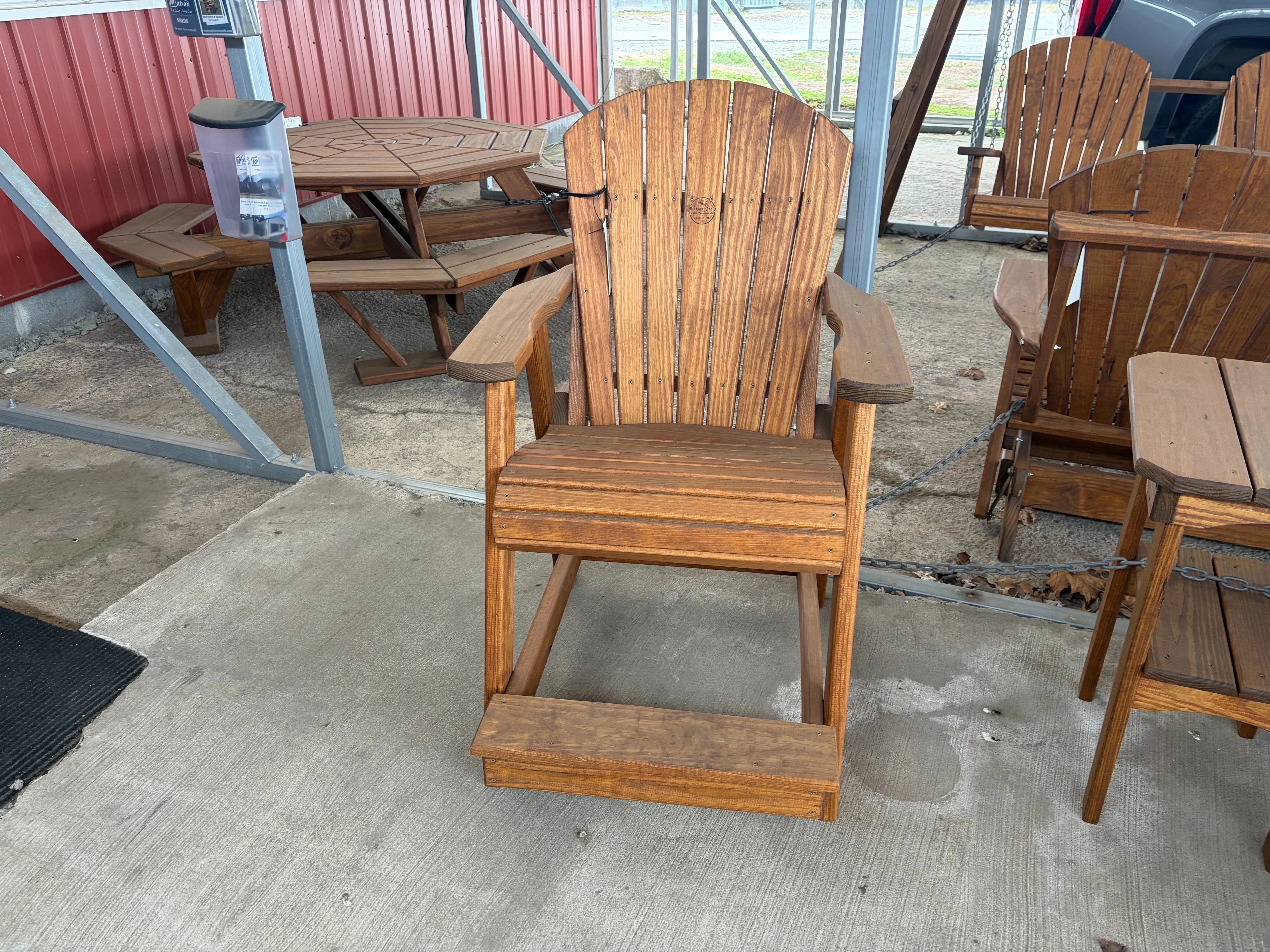 a wood stained and sealed chair sitting on concrete. It has arm rests and foot rests. It is chained up to other furniture