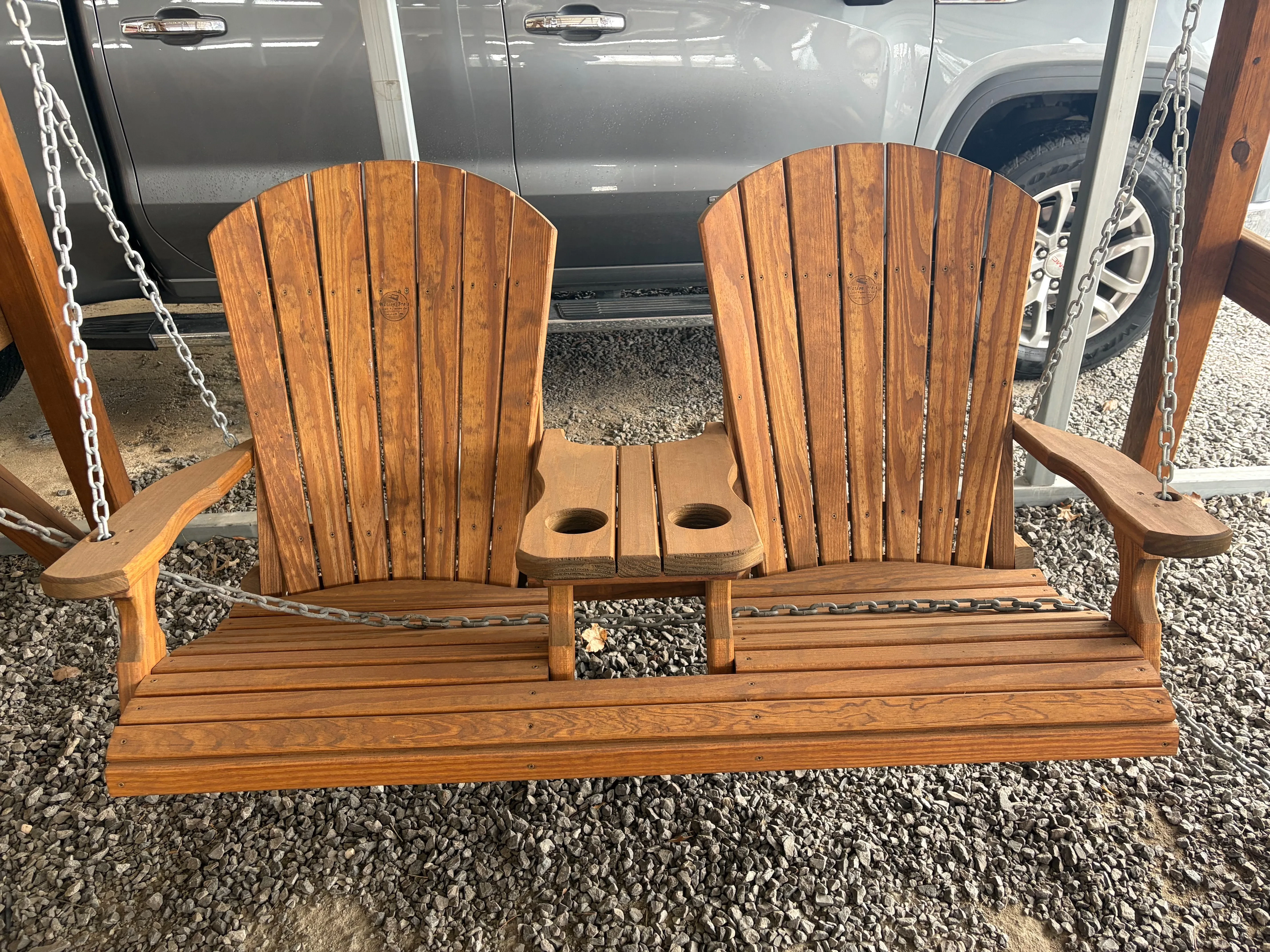 the front view of a wood stained and sealed bench swing. It has a center console with cupholders and is hovering over gravel