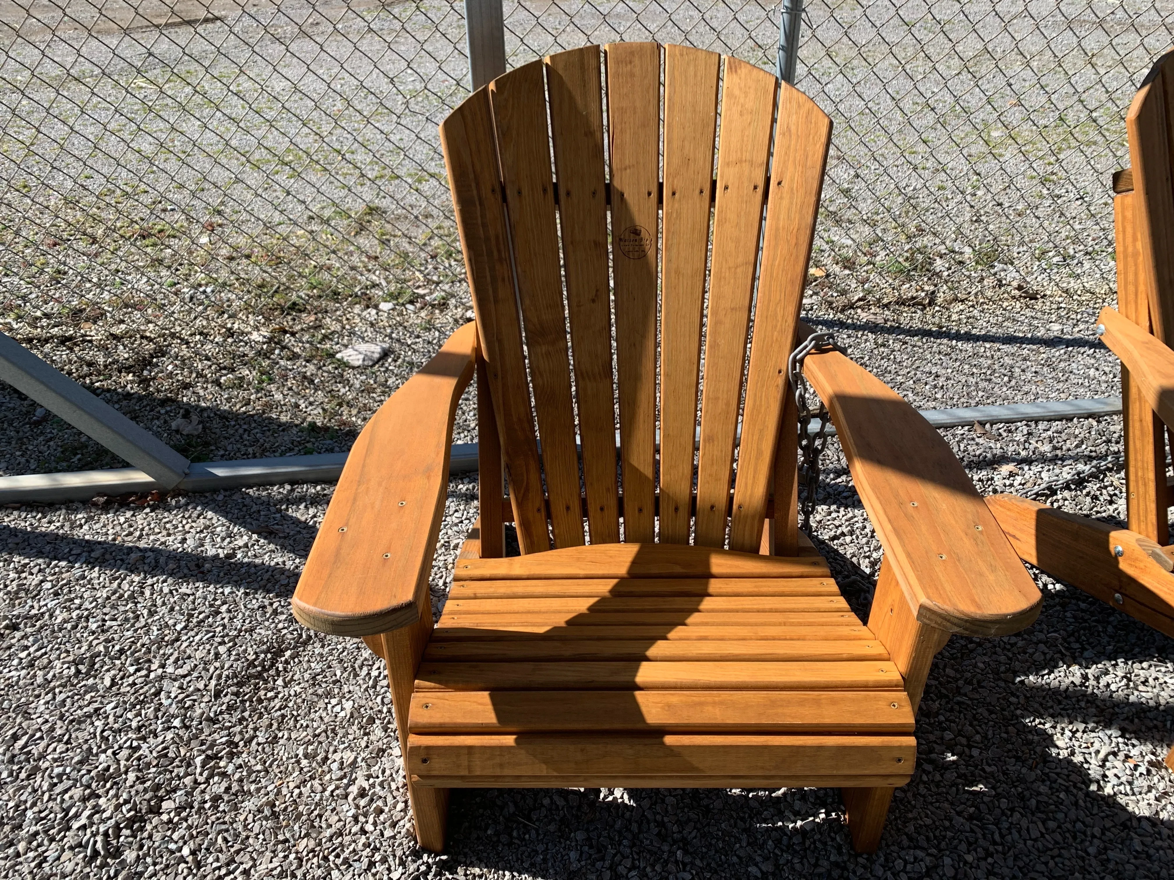 a wood stained and sealed chair sitting on gravel. There is a chain link fence behind it.