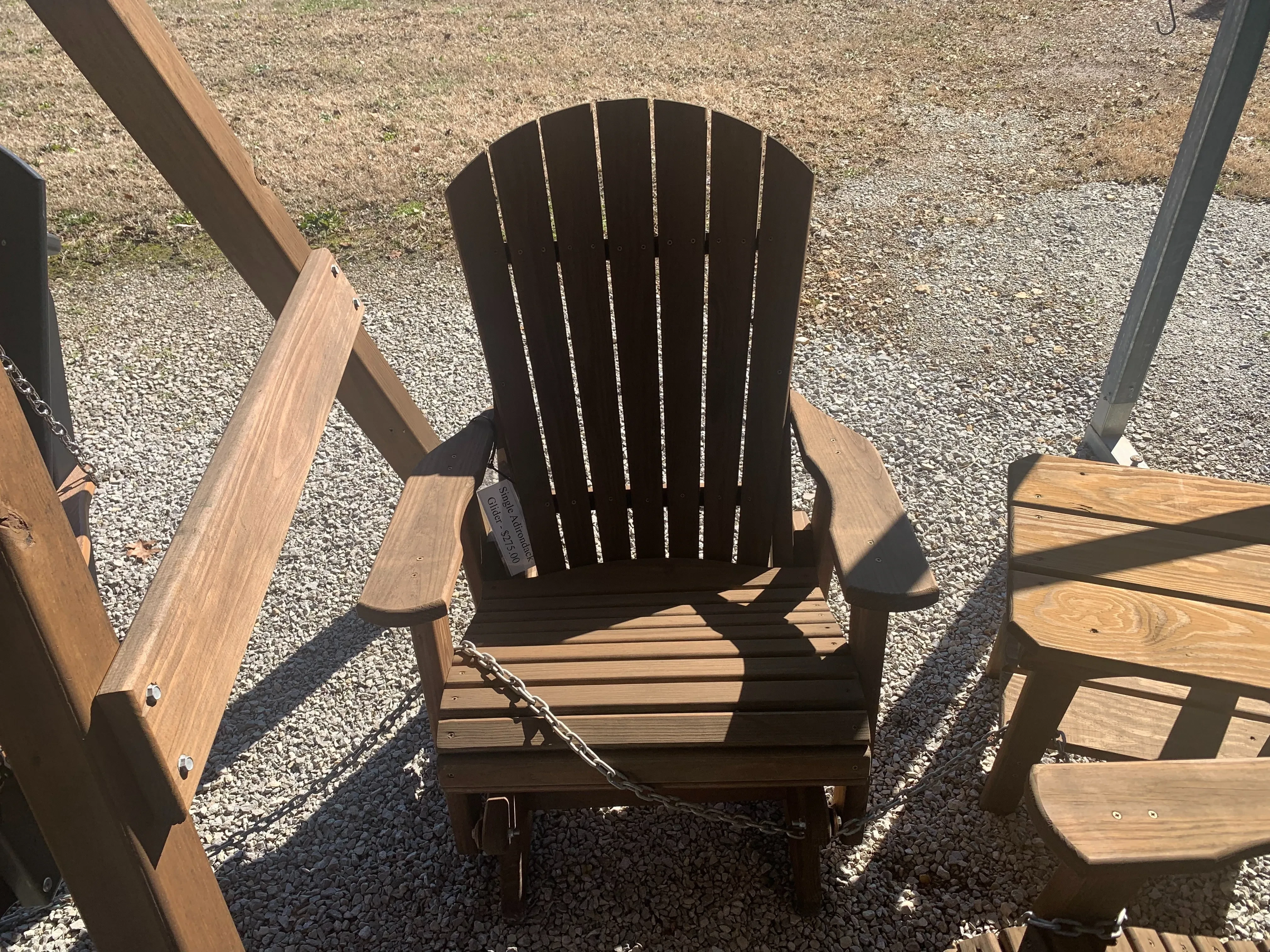 a wood stained and sealed chair sitting in the gravel. It has armrests and a white price tag. it can glide back and forth