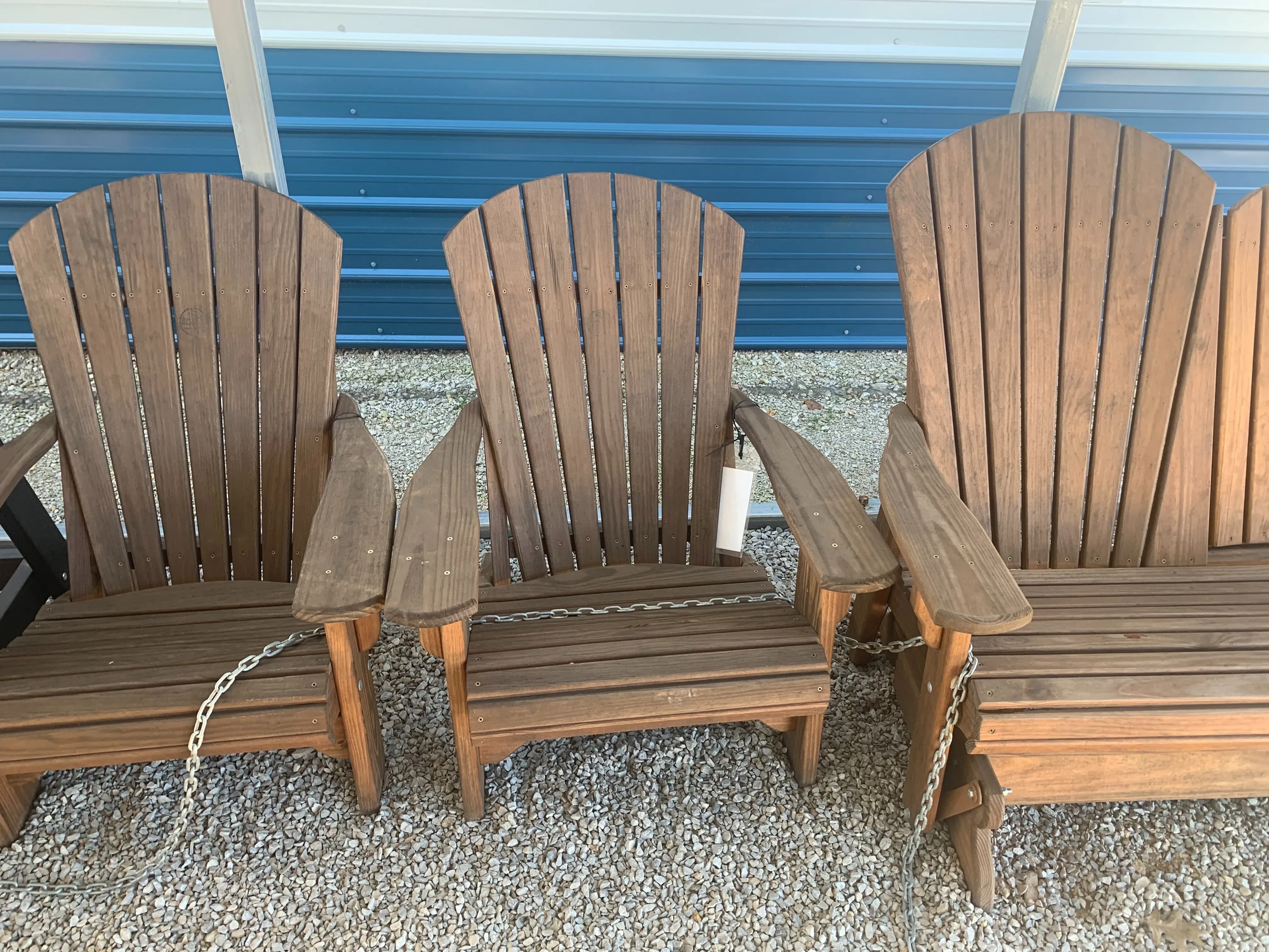 a wood stained and sealed chair sitting in the gravel. It is sitting next to other similar chairs. It has a white price tag