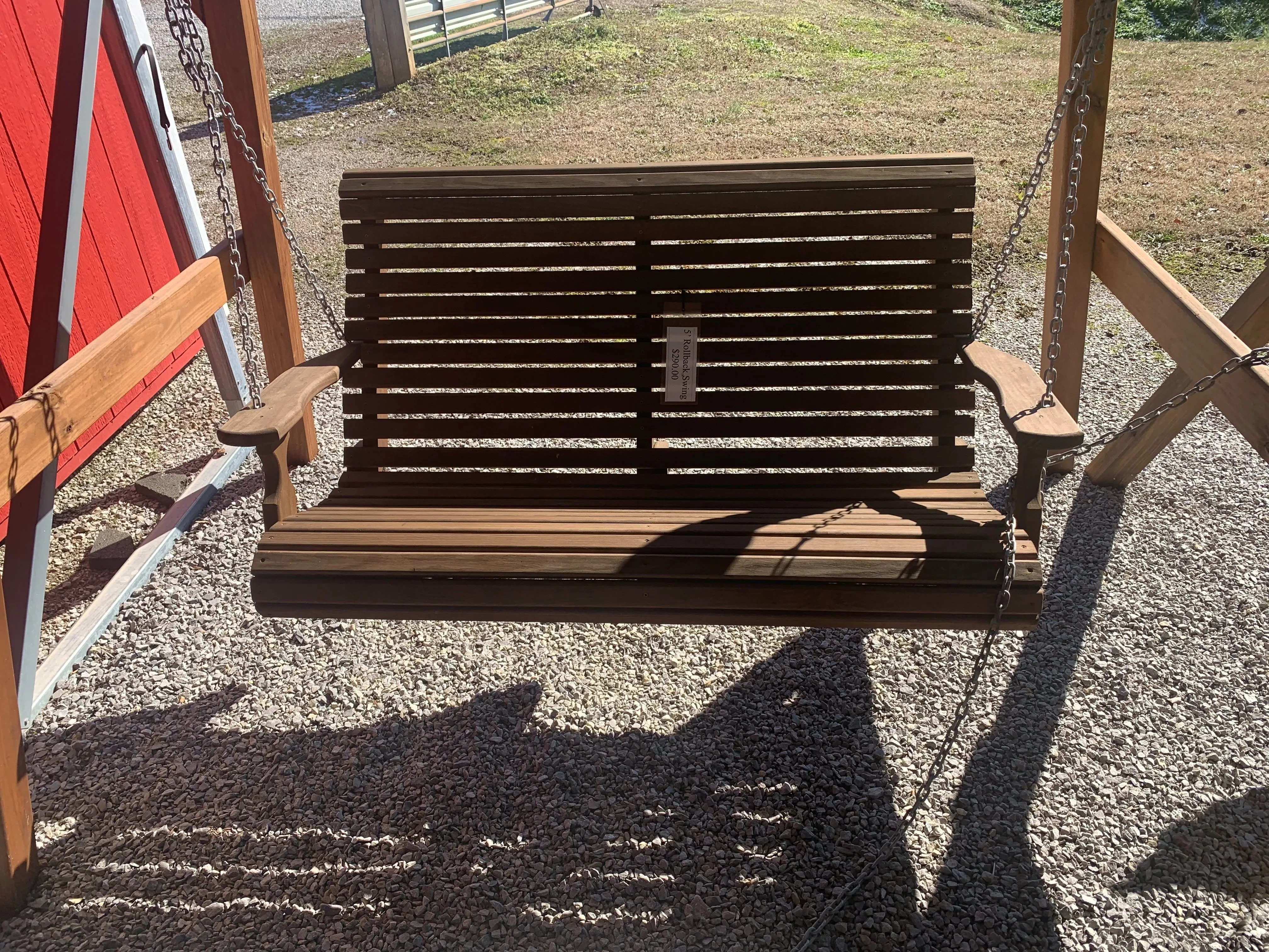 a wood stained and sealed bench swing hovering over gravel. It has a white price tag on the front