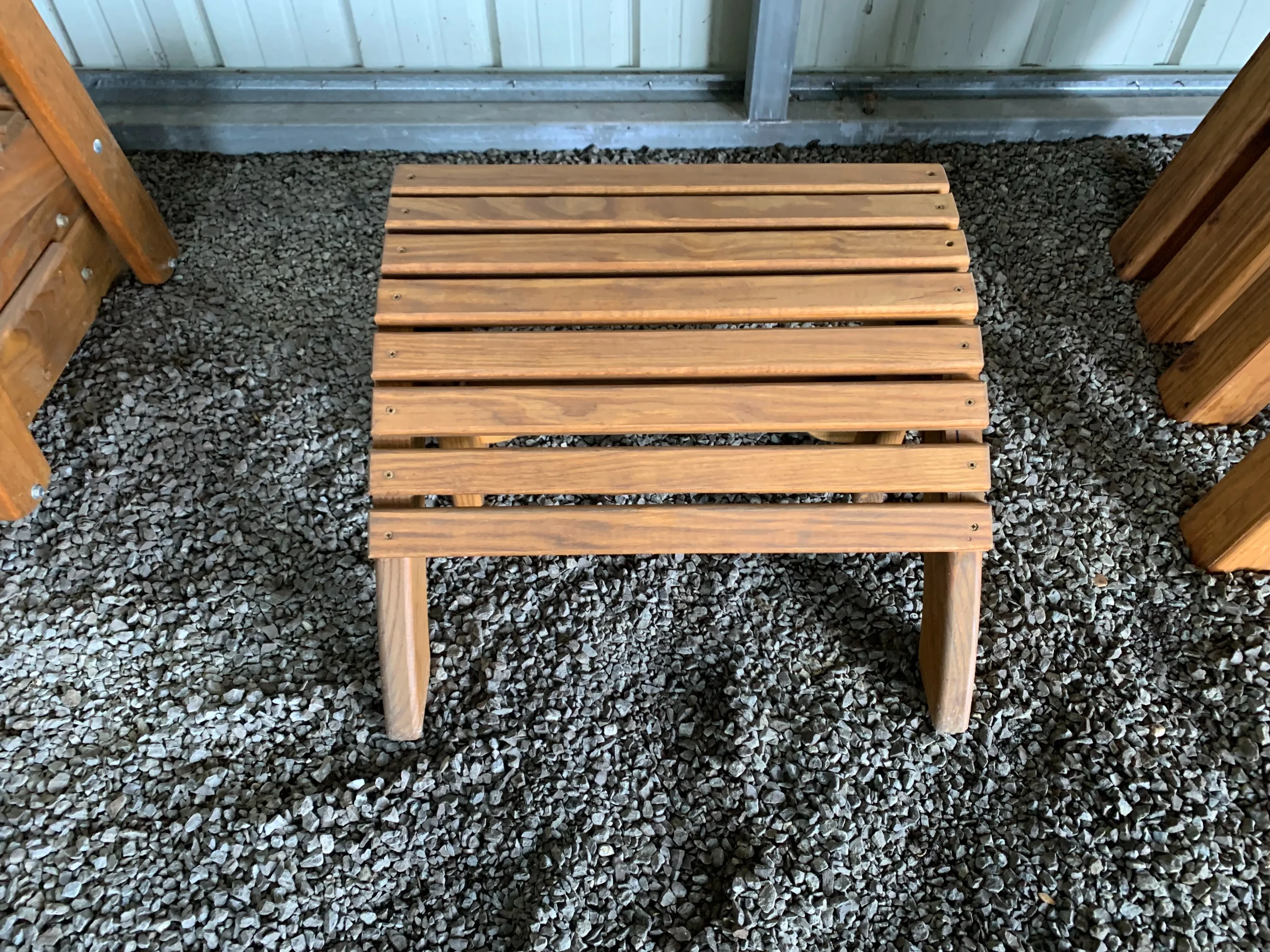 a wood footstool that is stained and sealed. It is sitting in the gravel. You can see other furniture nearby.