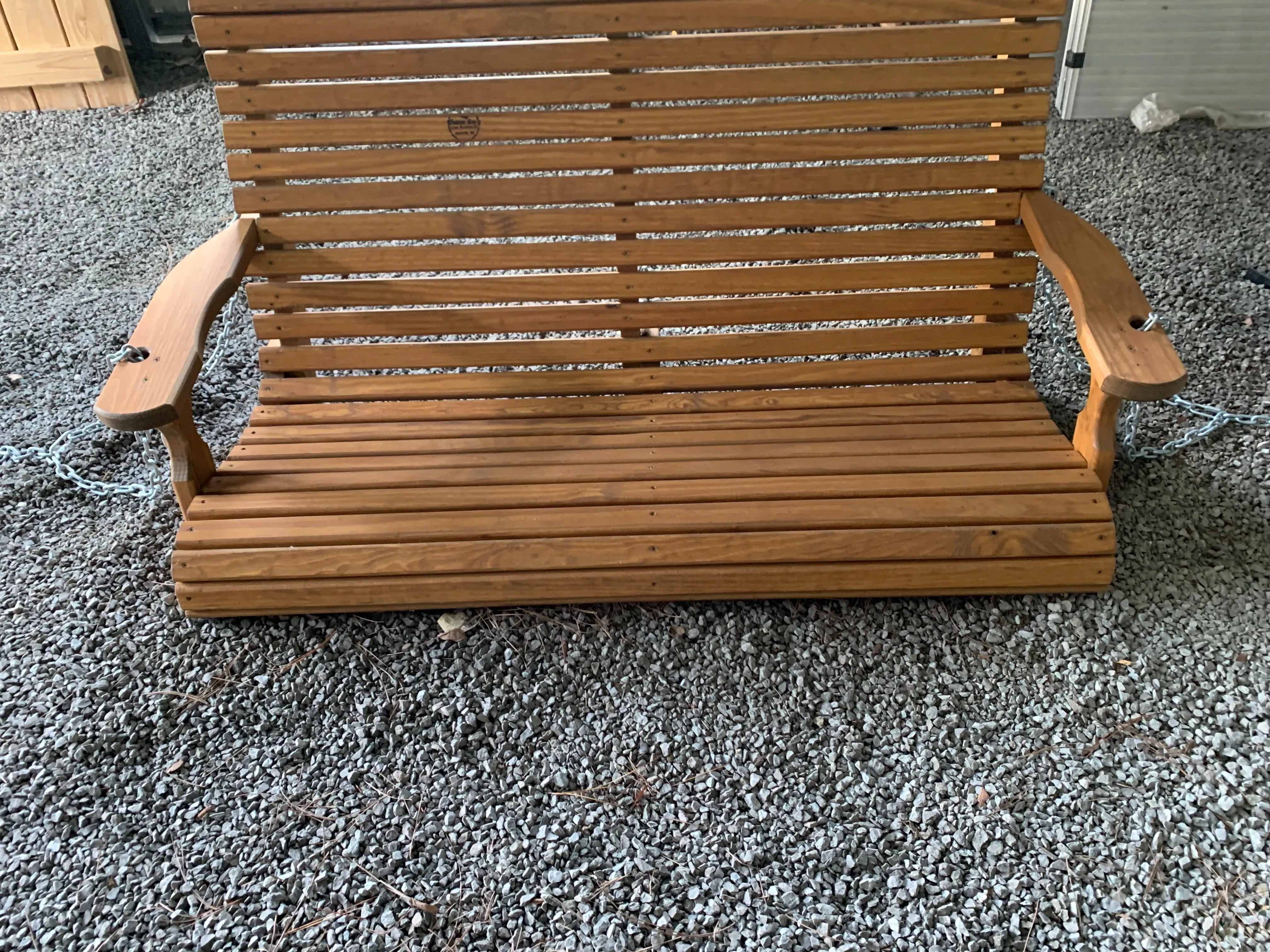 a swing bench sitting in the gravel. It is stained and sealed. It has slats that run horizontal