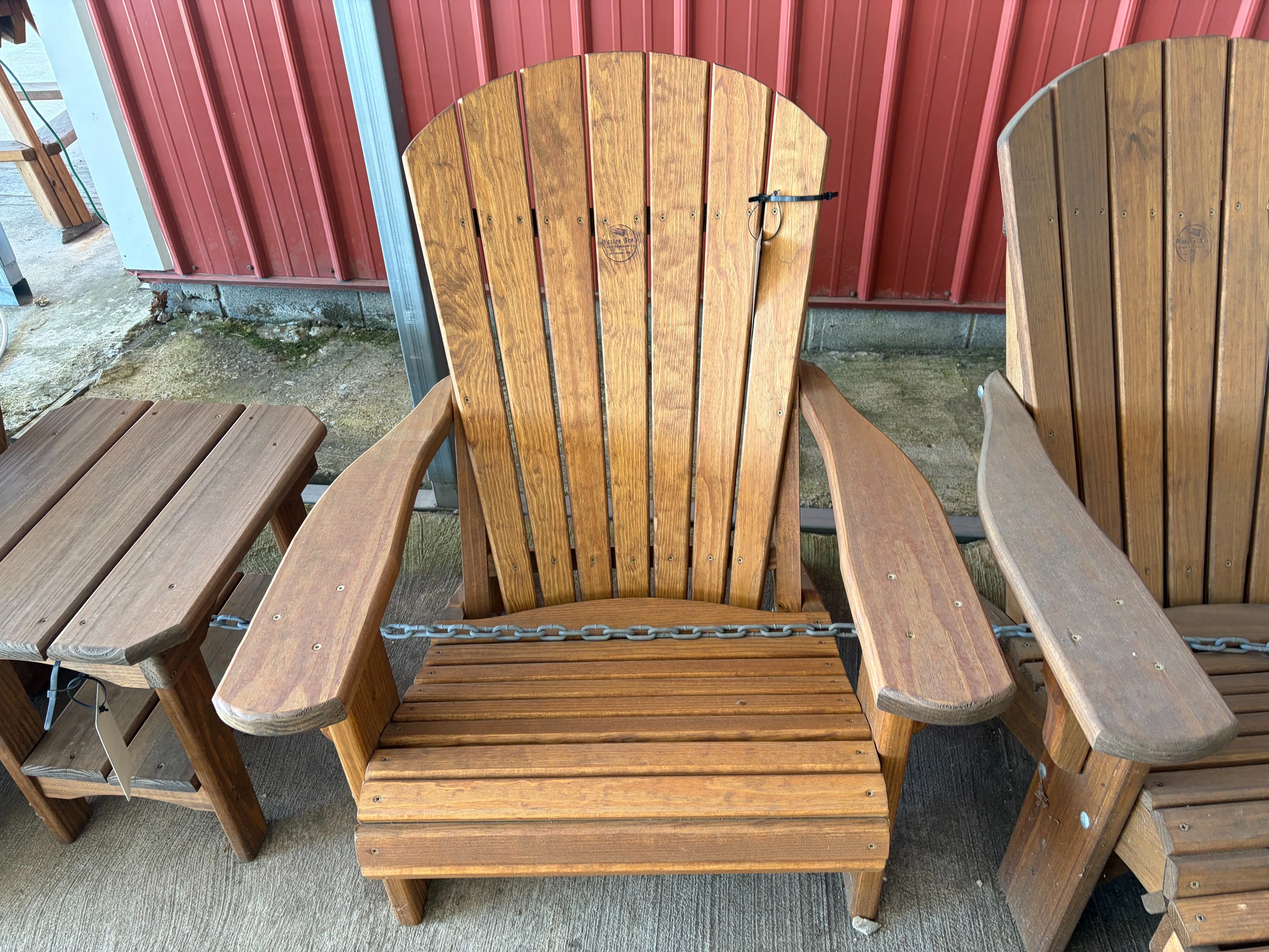 a wood stained and sealed chair that is sitting on concrete next to other furniture. It is chained up to the other furniture