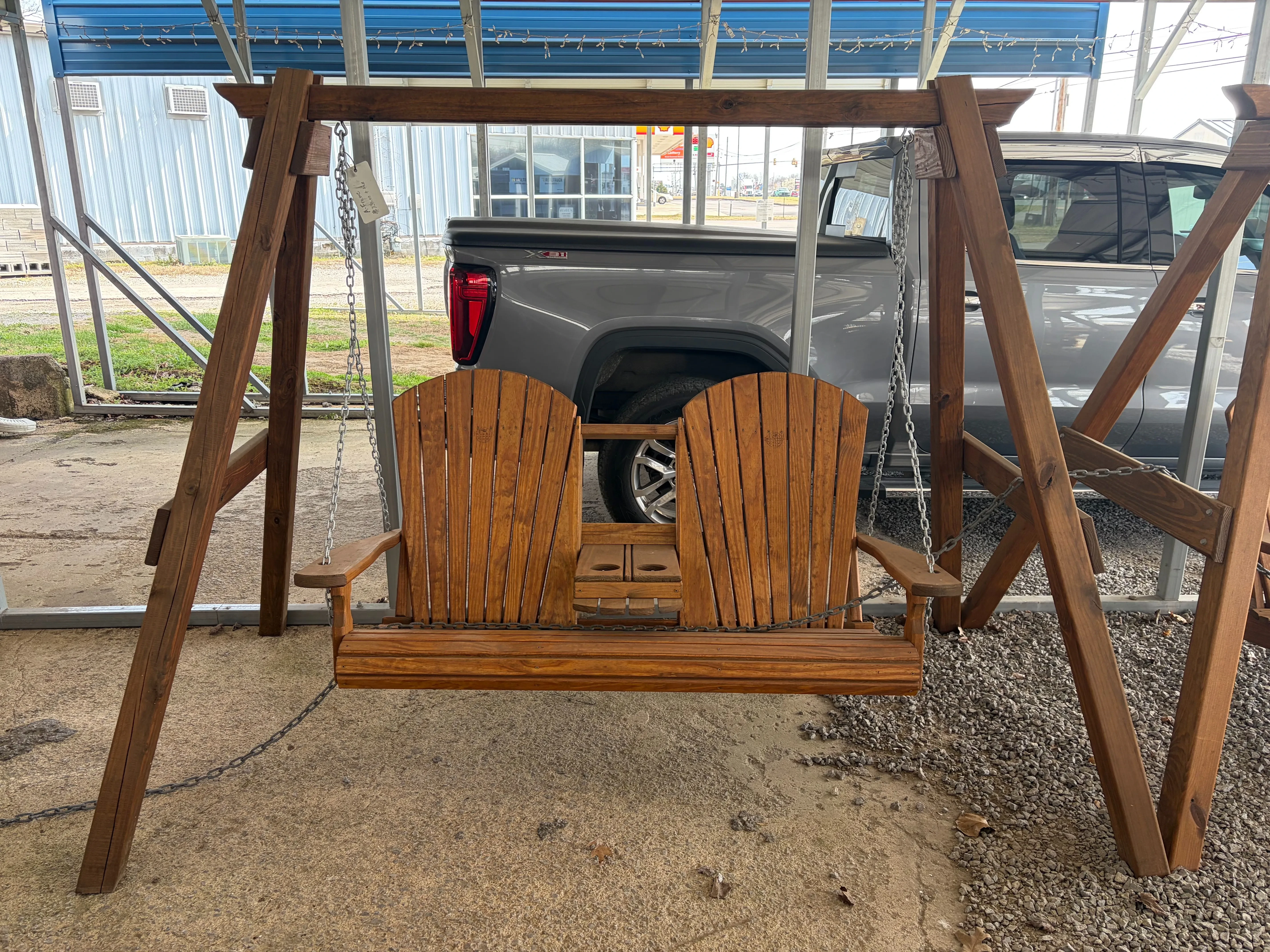a wooden a-frame structure holding up a wood bench swing by chains. It is sitting on concrete. There is a truck behind it