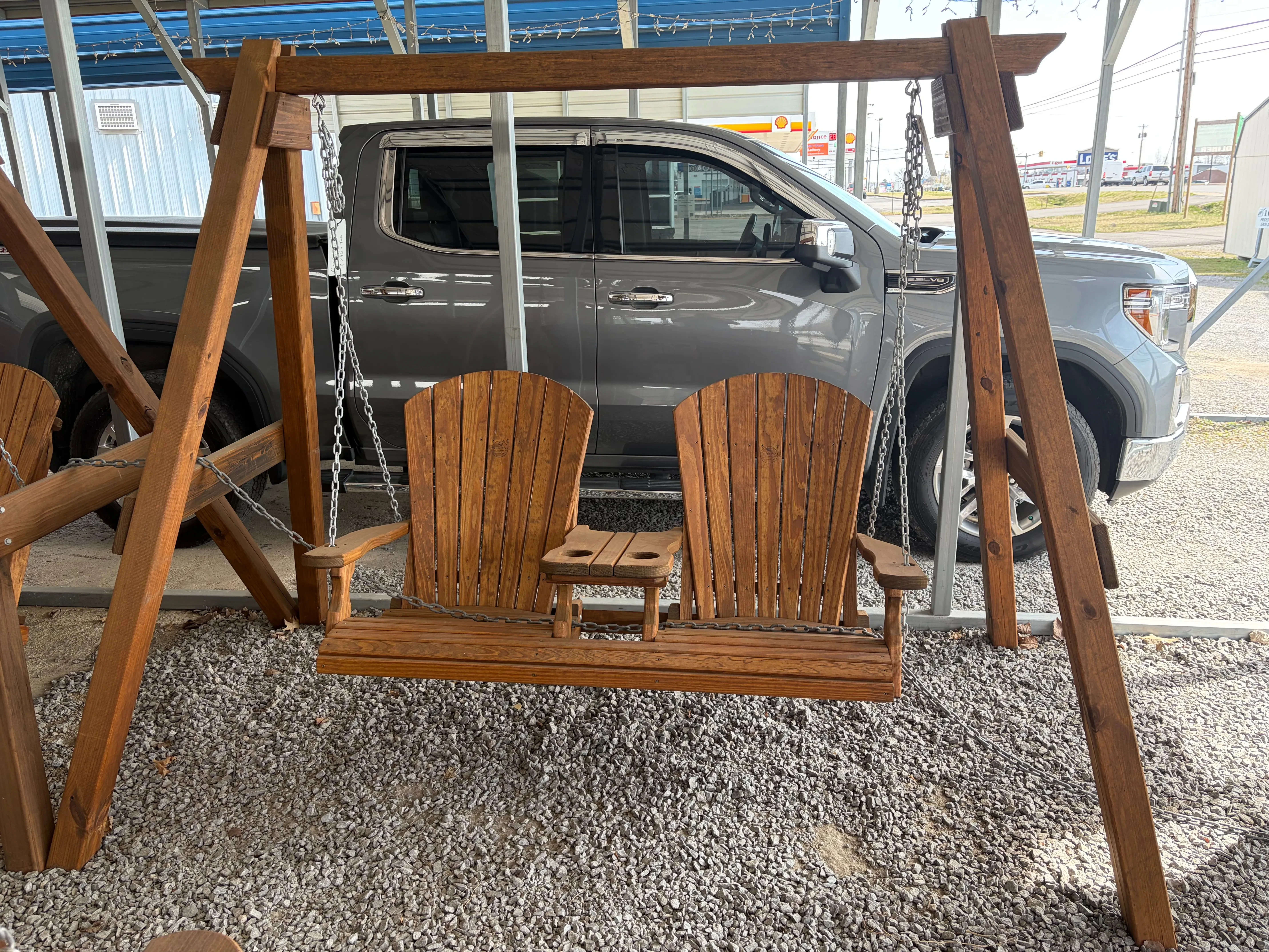 a stained and sealed wood bench swing attached to a wooden a-frame structure. It is hovering over gravel in front of a truck
