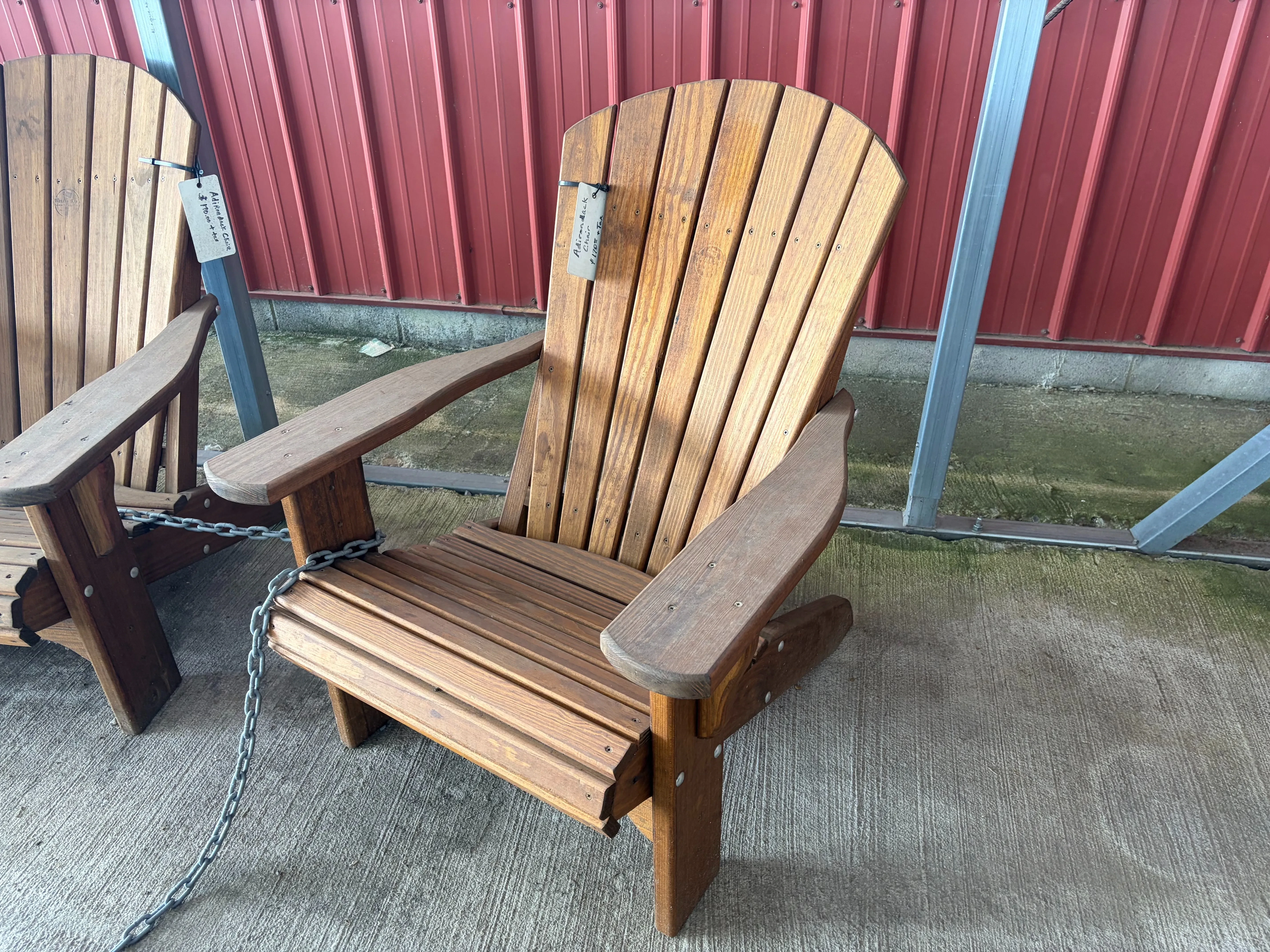 a wood stained and sealed chair sitting on concrete. It has a white price tag. It is chained up to other nearby furniture