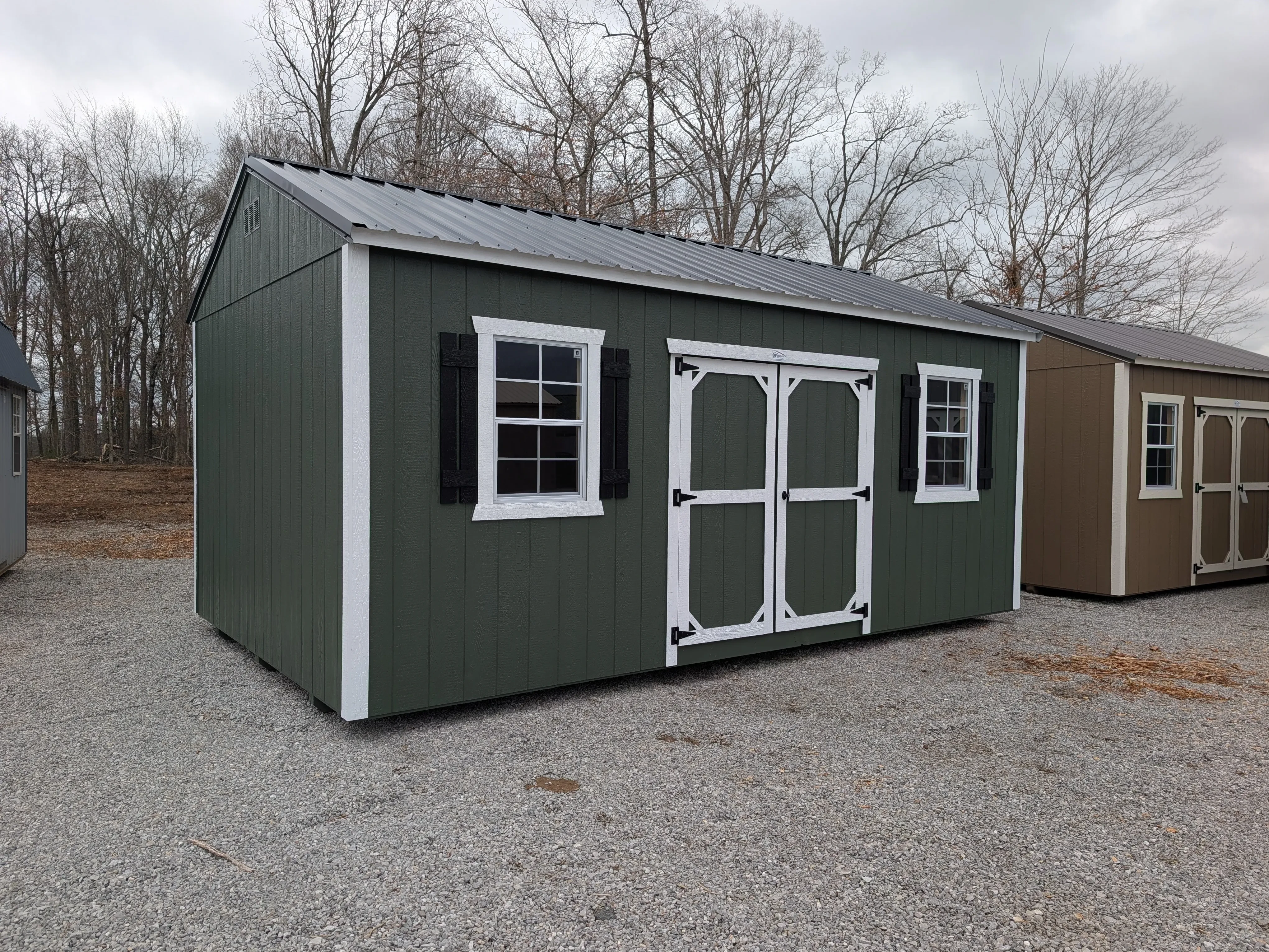 a green garden shed sitting on gravel. It has double doors and two windows. The windows have shutters. The trim is white