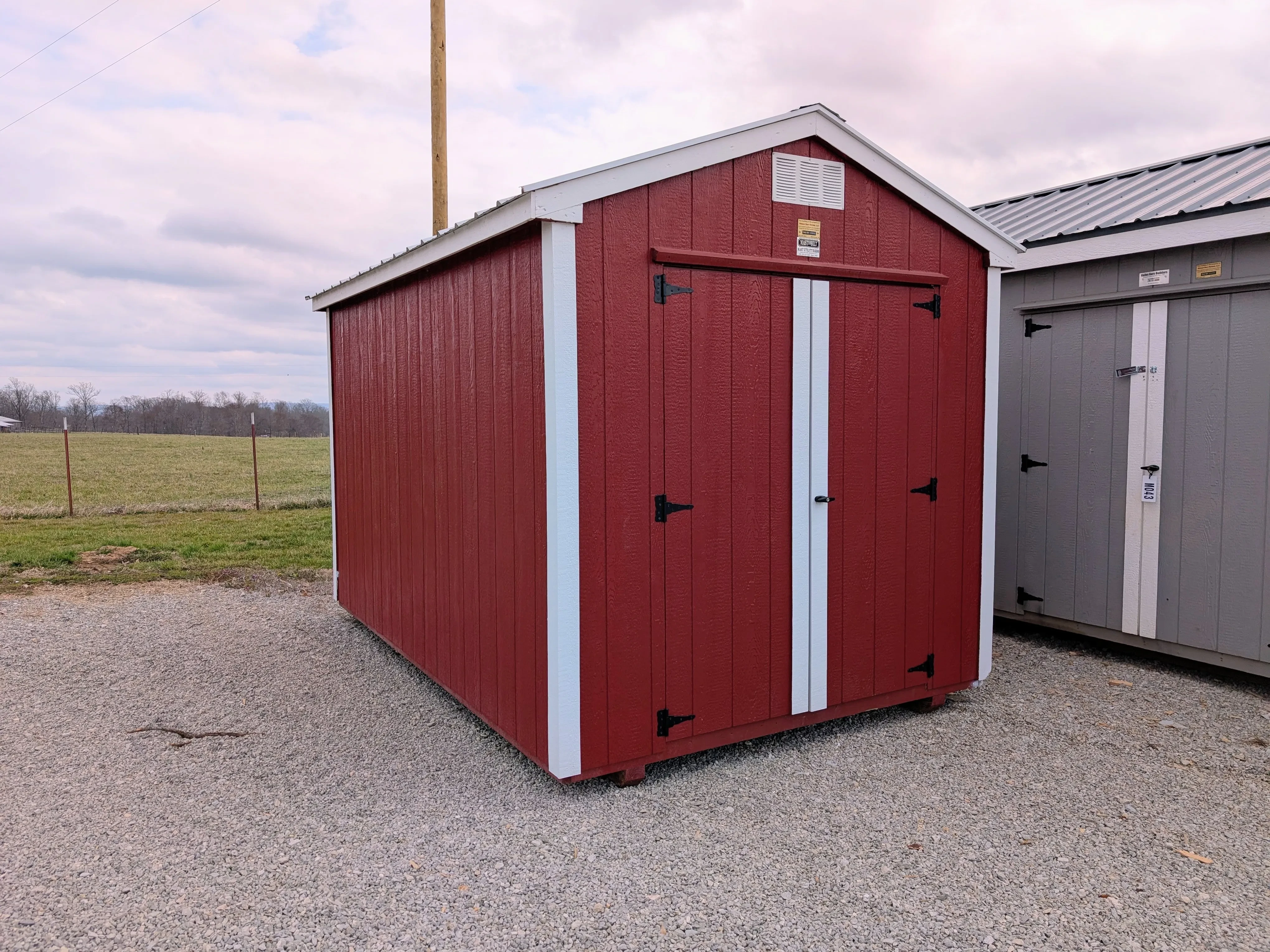 a small red shed with white trim sitting on gravel. There is a field behind it and another gray shed next to it