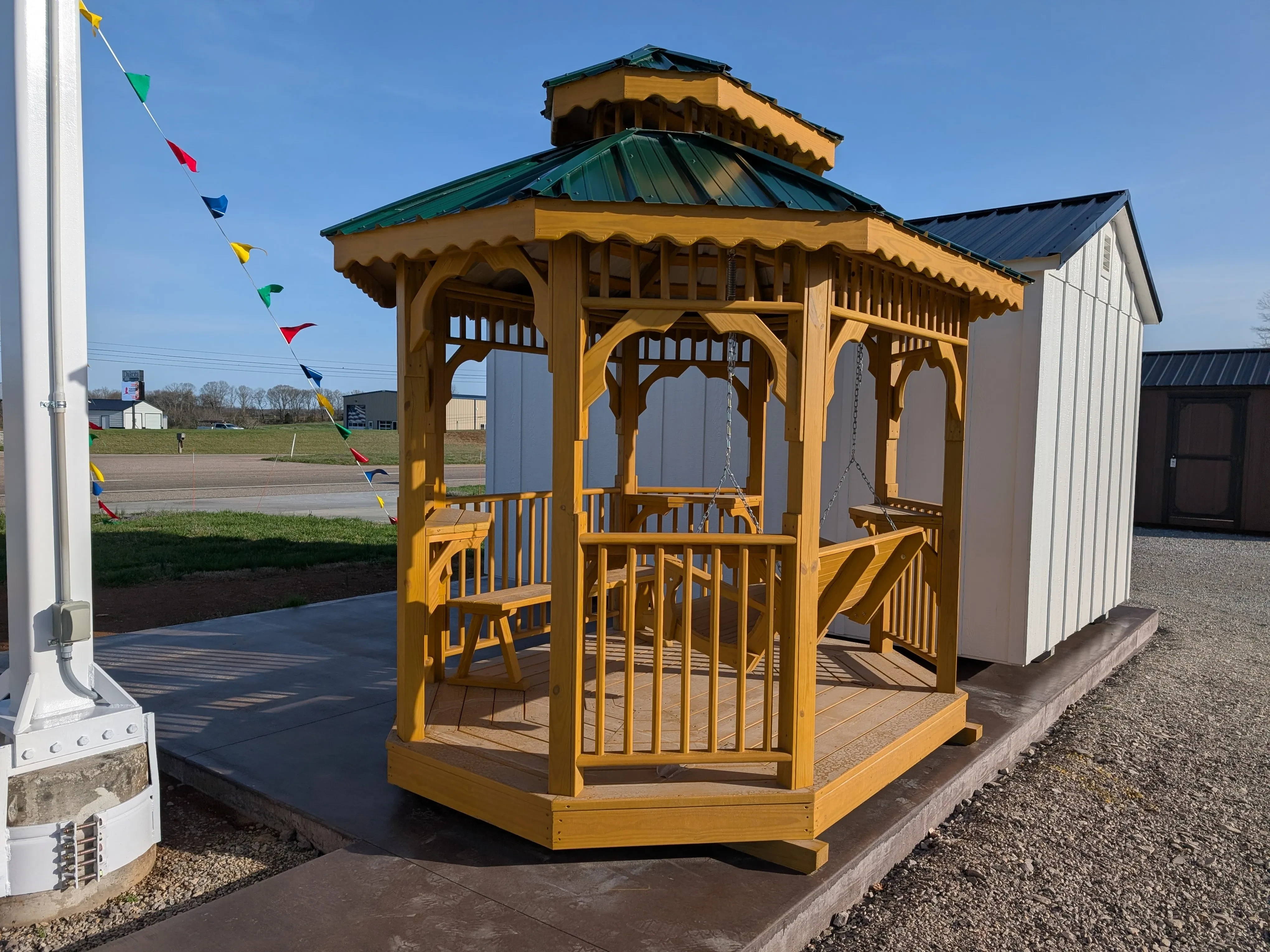 a honey gold stained colored teahouse with a double roof sitting on a concrete pad. There is a white shed behind it