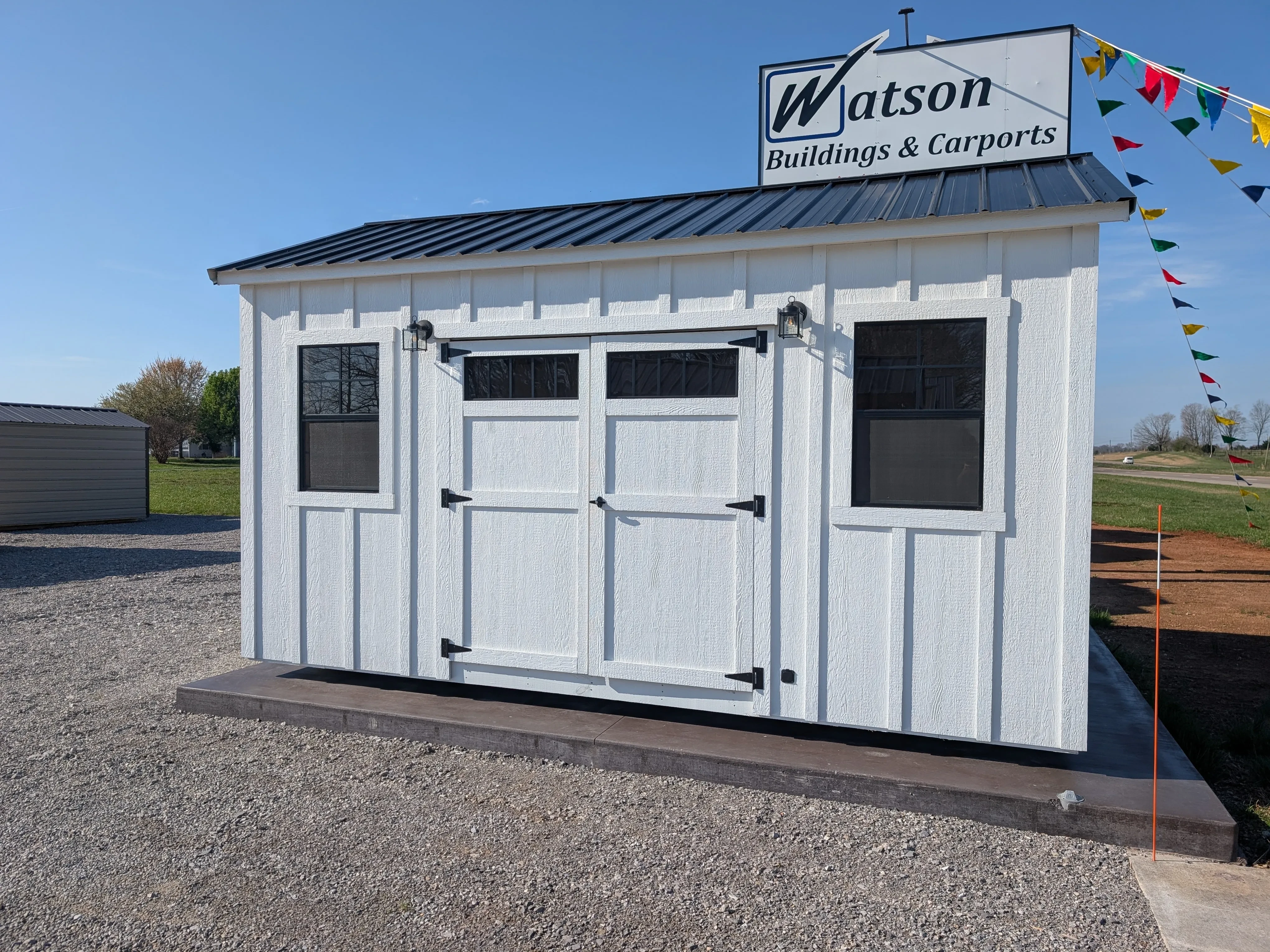 a white board & batten shed with black windows and outside lights. It has a black metal roof and is near other sheds.