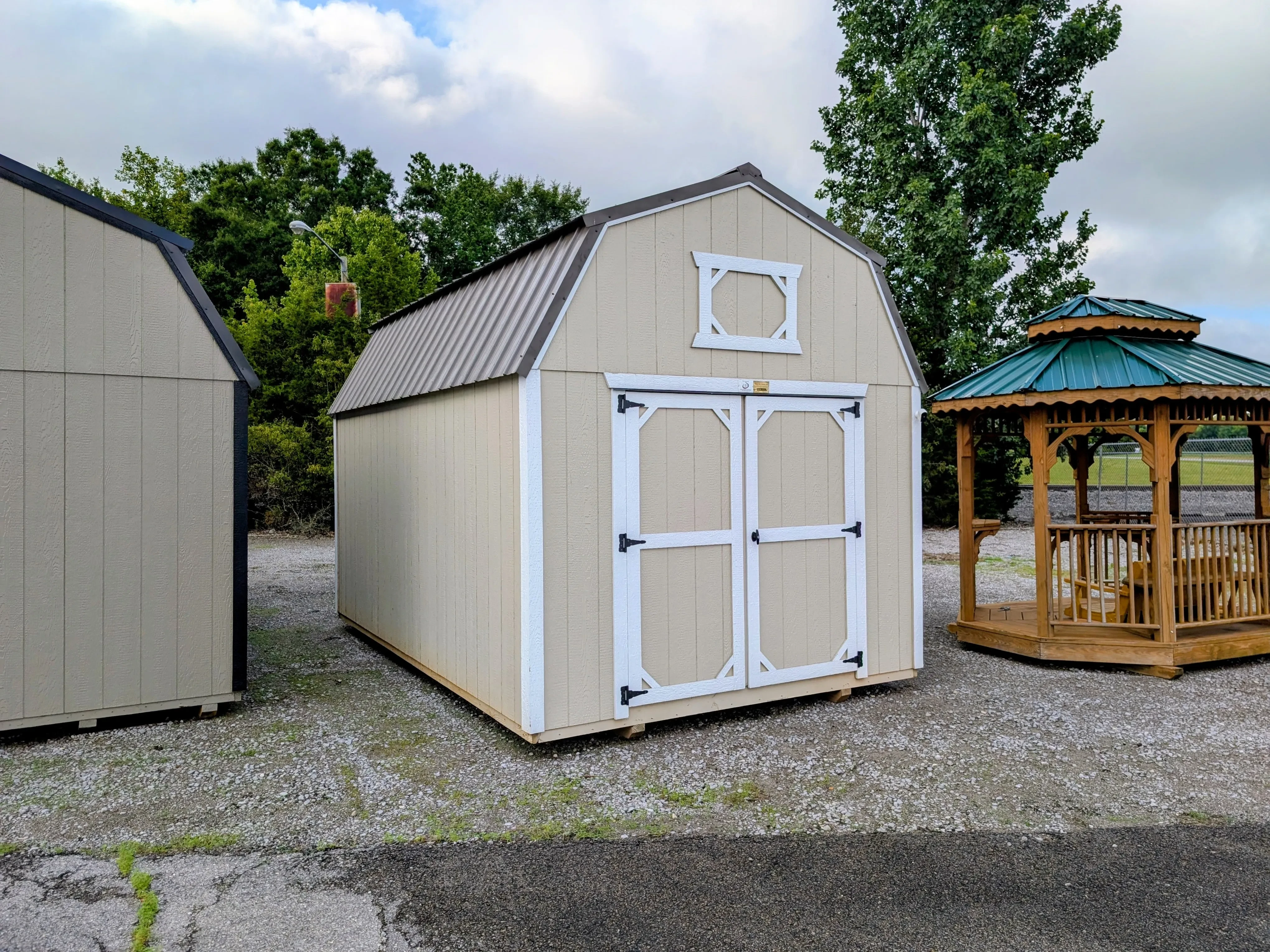 A light stone colored lofted barn with double doors in the front and white trim. It is next to a wood gazebo.