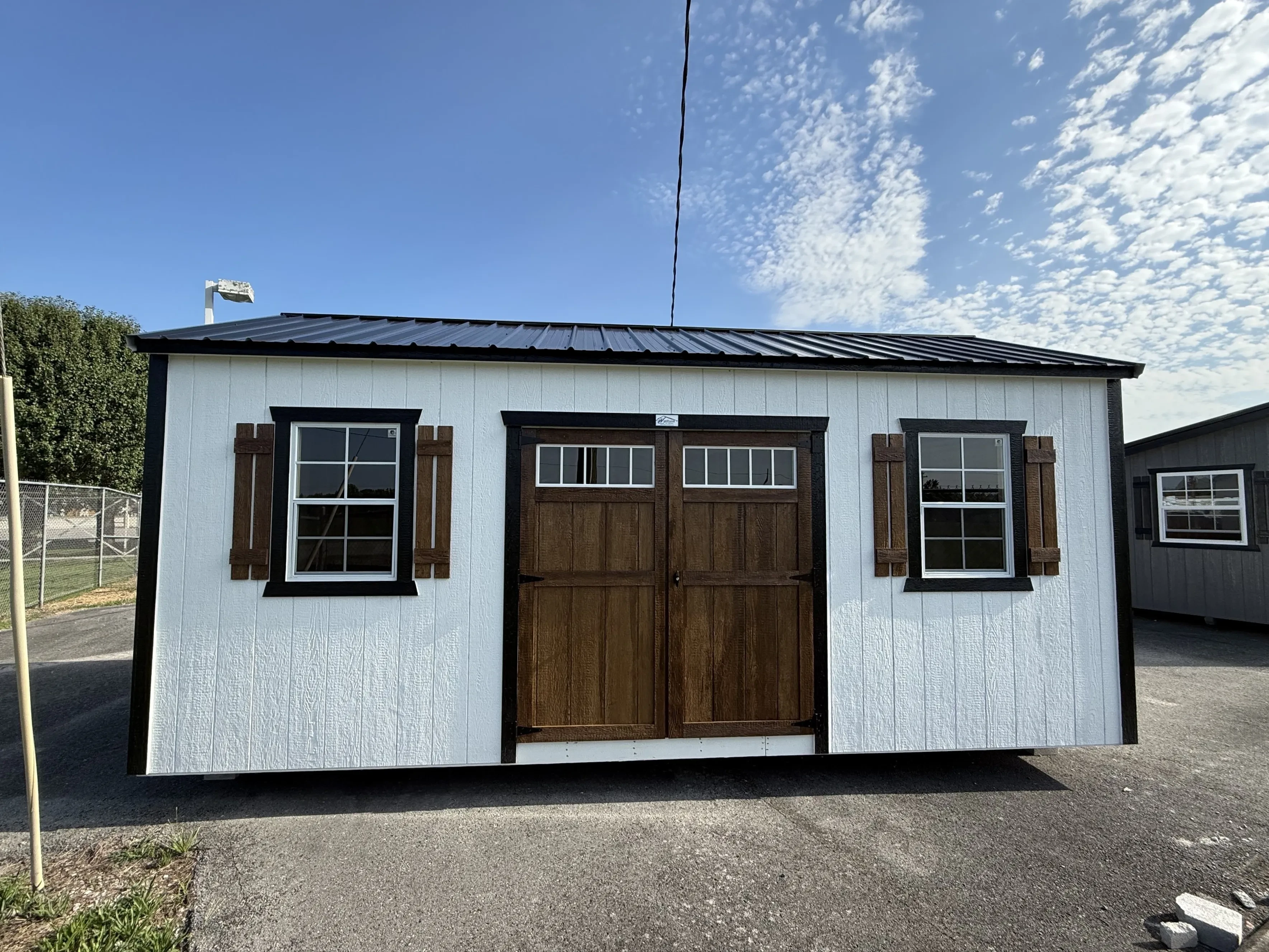 a white garden shed with a black metal roof and two windows. It has wood stained doors and shutters.